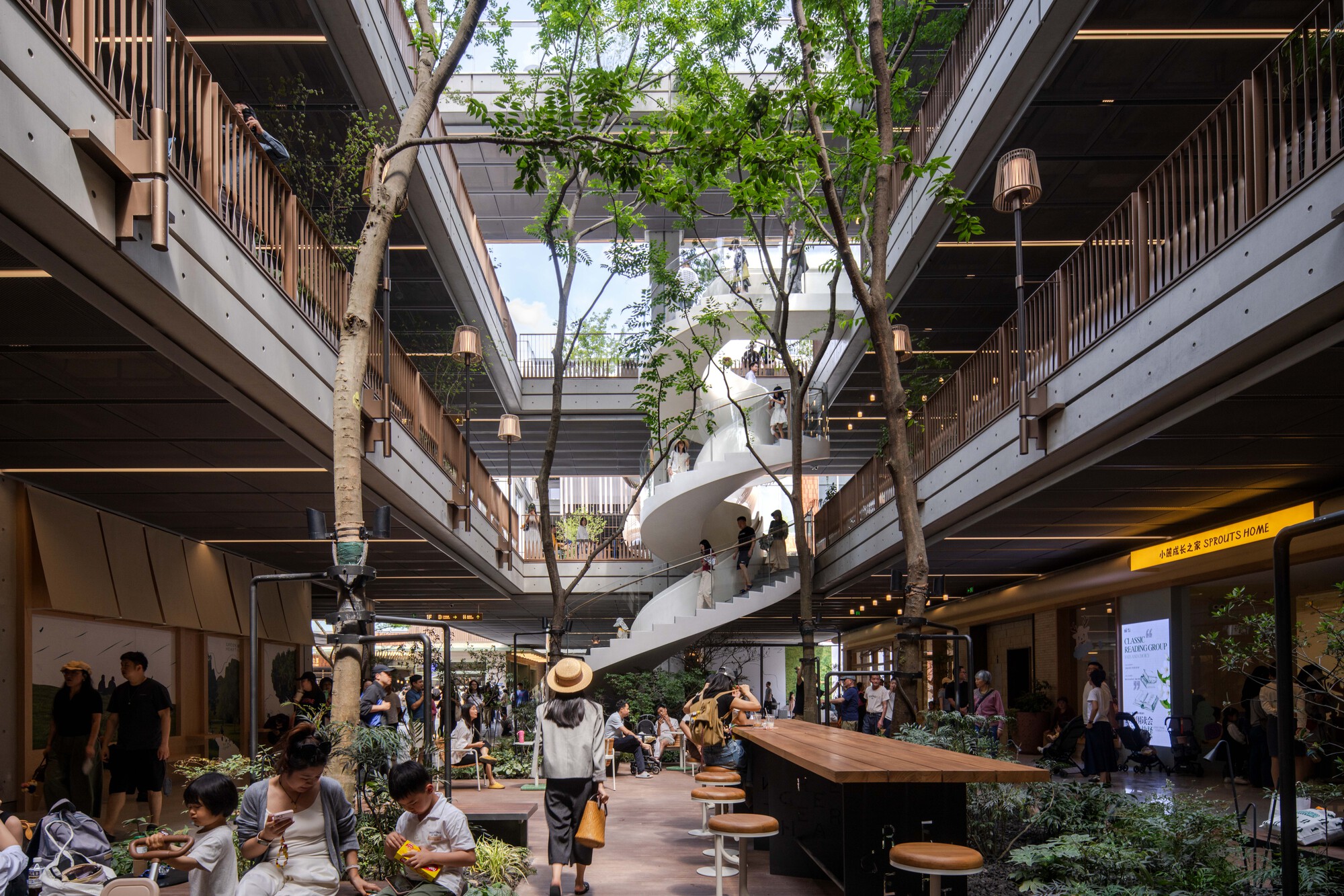 Crowded atrium court with a white spiral stair, timber-railed balconies, a long wooden counter and trees rising between the levels