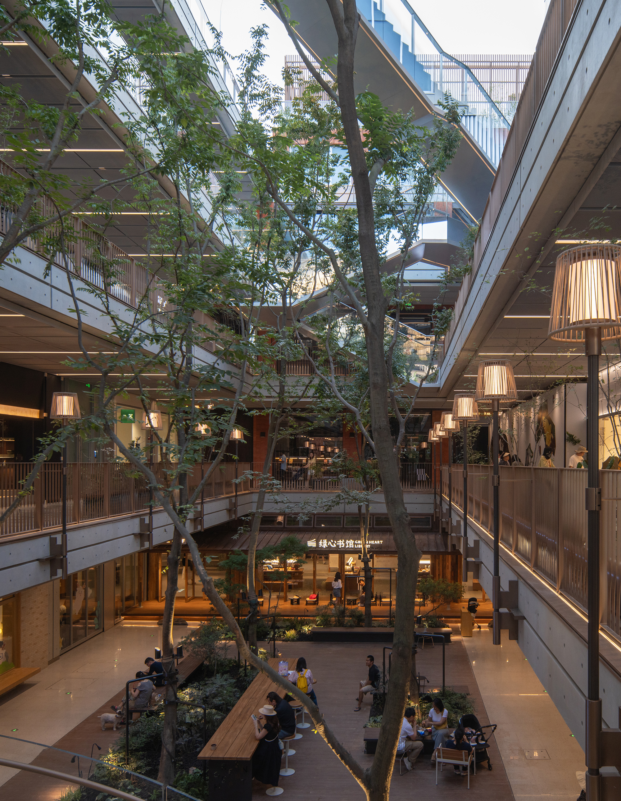 Vertical view down a tree-filled atrium with timber-screened balconies, lantern-shaped lamps and people seated at a long wooden table in evening light
