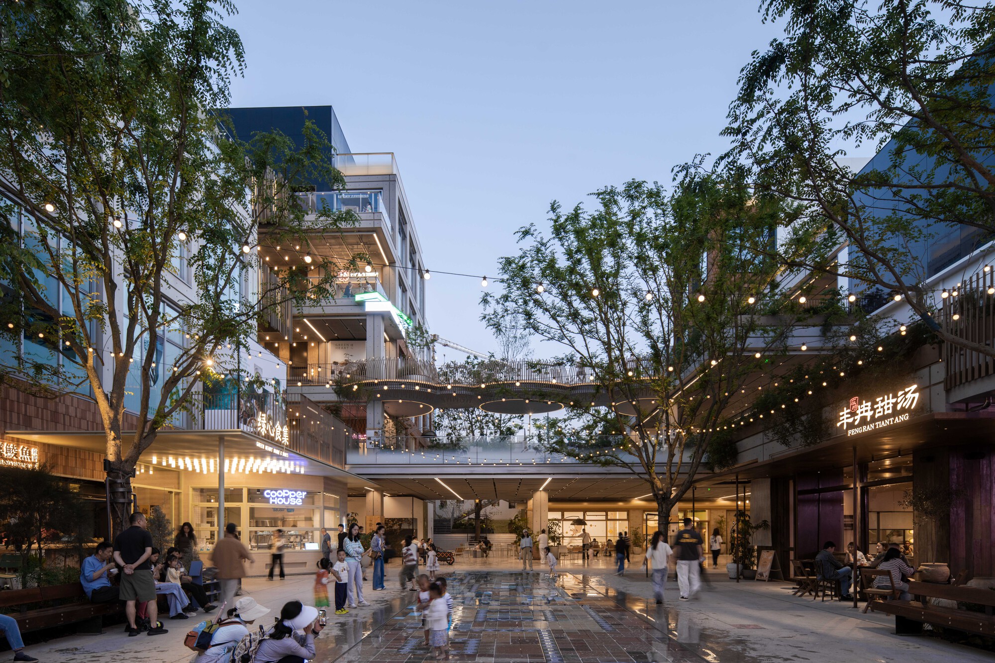 Pedestrian plaza with wet paving, string lights and shopfronts, with visitors gathered beneath trees at dusk