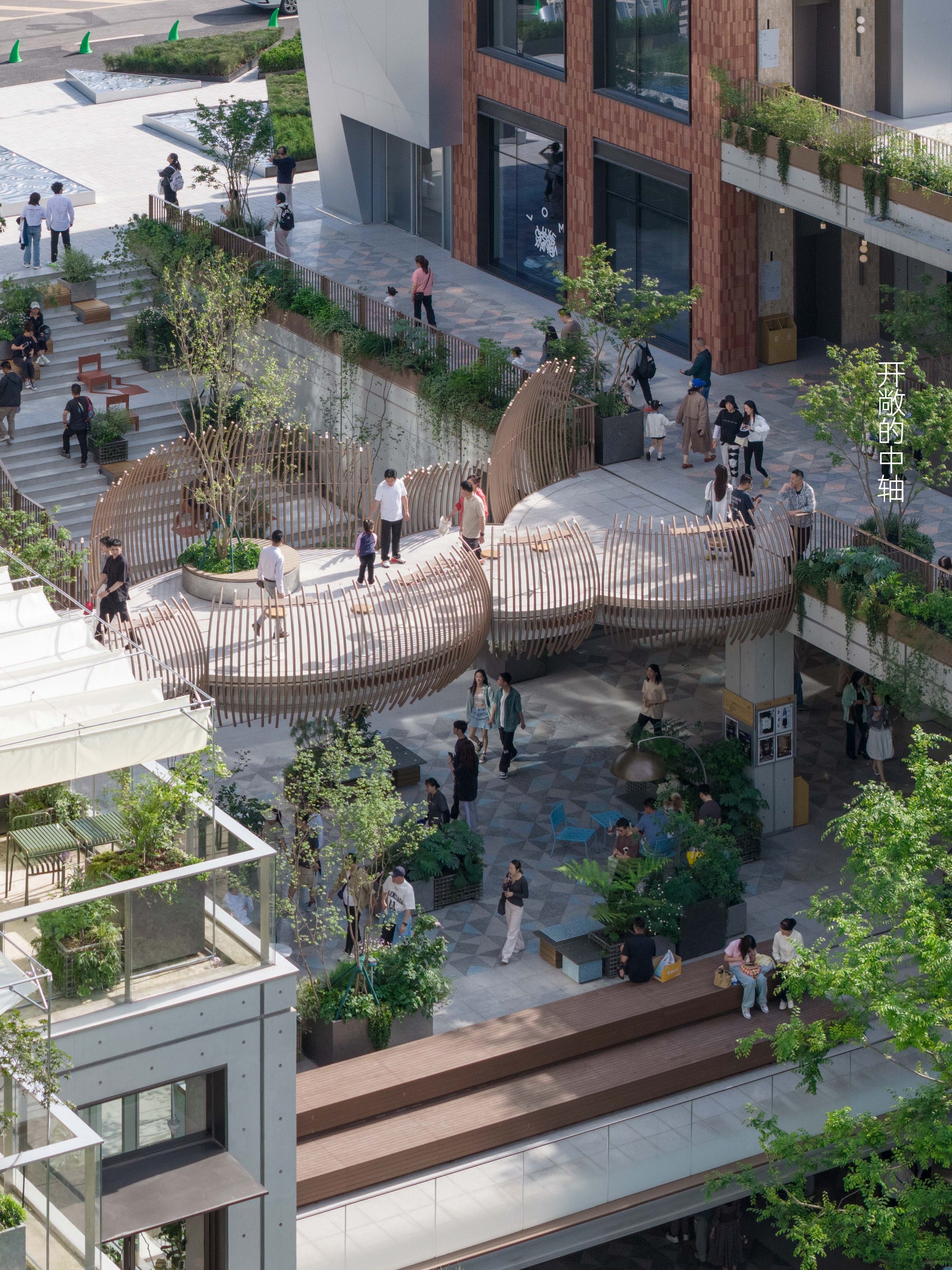 Aerial view of a slatted timber pavilion with curved edges over a terraced plaza, with people walking among planters