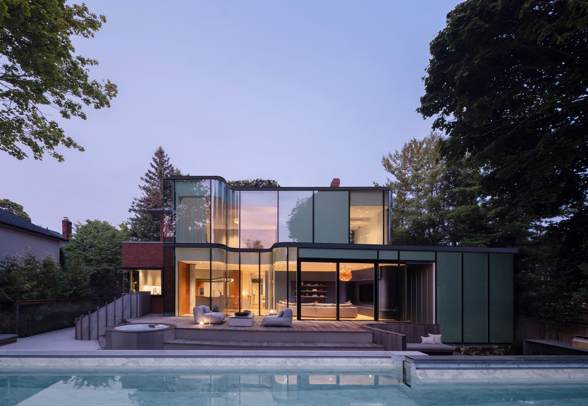 Rear elevation of the curved glass addition with green panels, lit interior and a swimming pool in the foreground at dusk