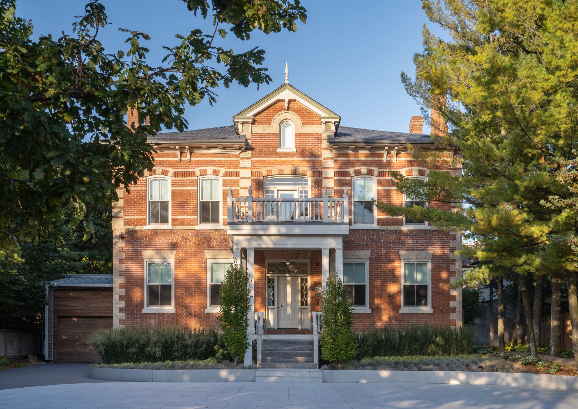 Front facade of the red brick house with white balcony, columned porch and pine tree in late afternoon sun