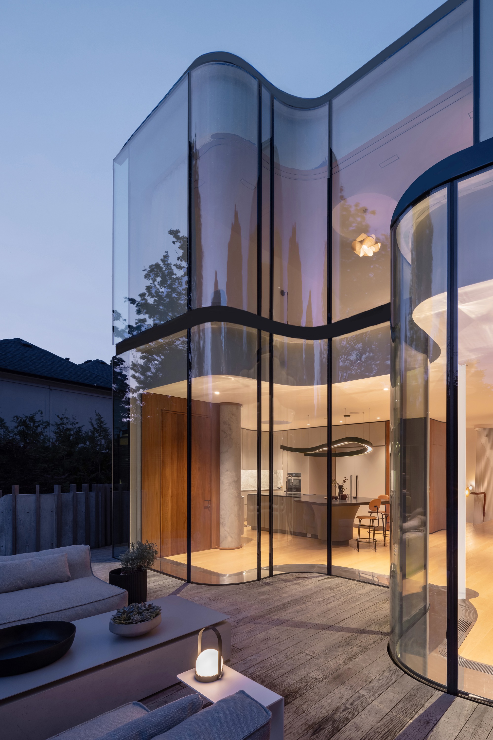 Close view of the undulating glass walls enclosing the kitchen, seen from a timber deck with lounge seating at dusk