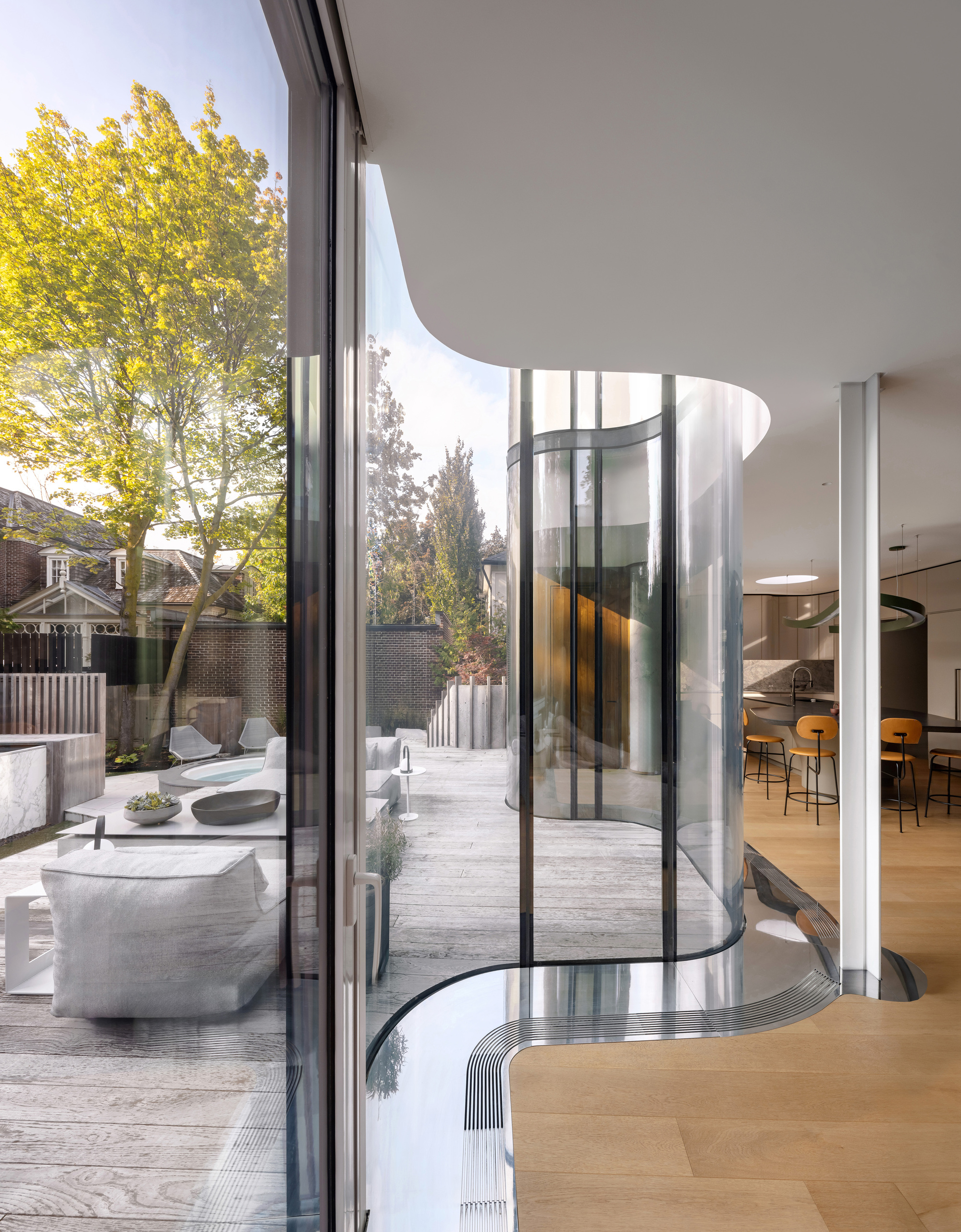 View from inside along the curved glass wall and floor edge toward the sunlit garden deck and yellow-leaved tree