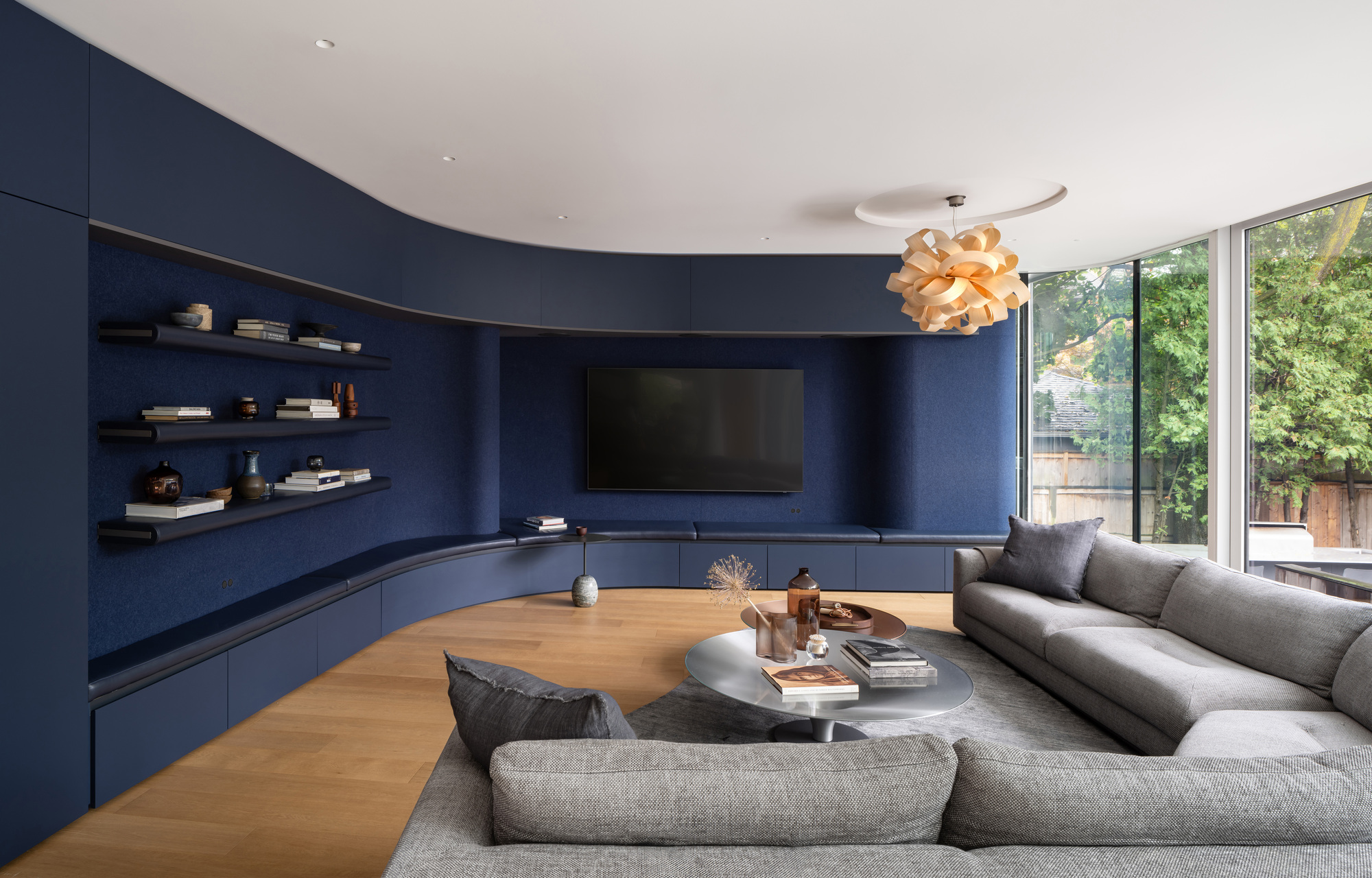 Living room with curved navy built-in shelving, wraparound grey sofa and wooden pendant lamp beside a glass corner