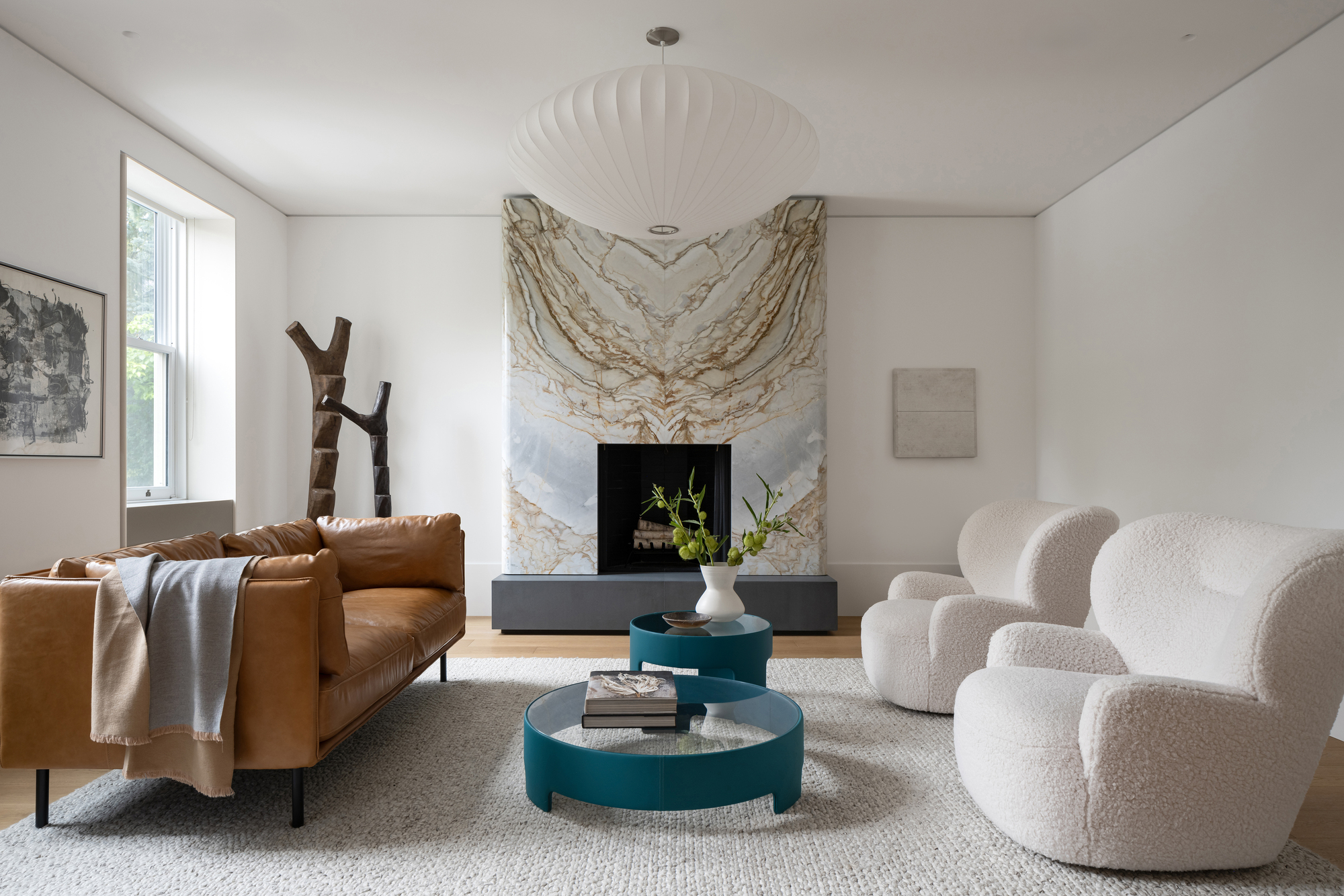 Sitting room with veined stone fireplace surround, tan leather sofa, boucle armchairs and a white pendant lamp in soft daylight