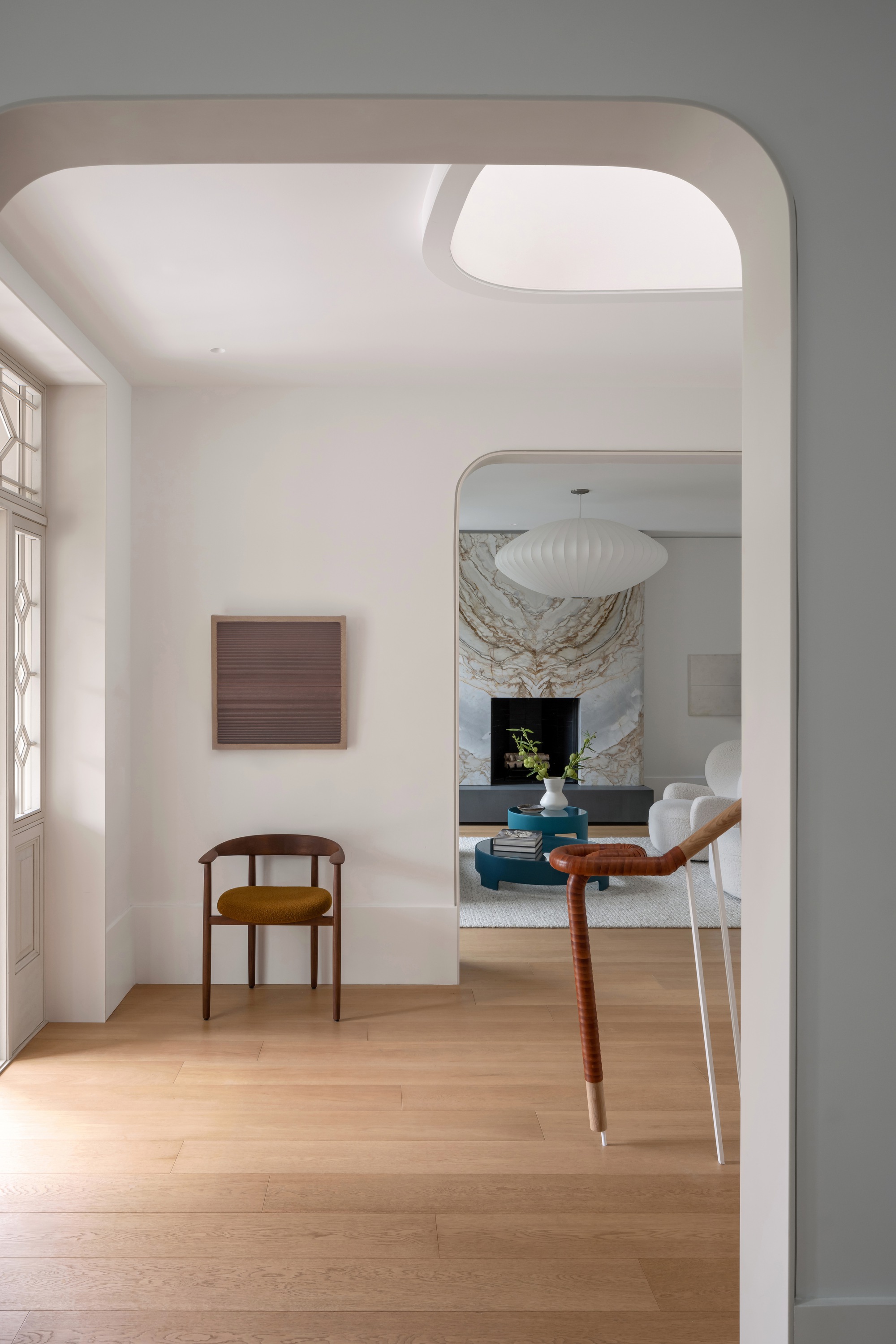 Rounded arched opening framing a wooden chair, framed artwork and a marble-backed fireplace beyond, lit by daylight from a skylight