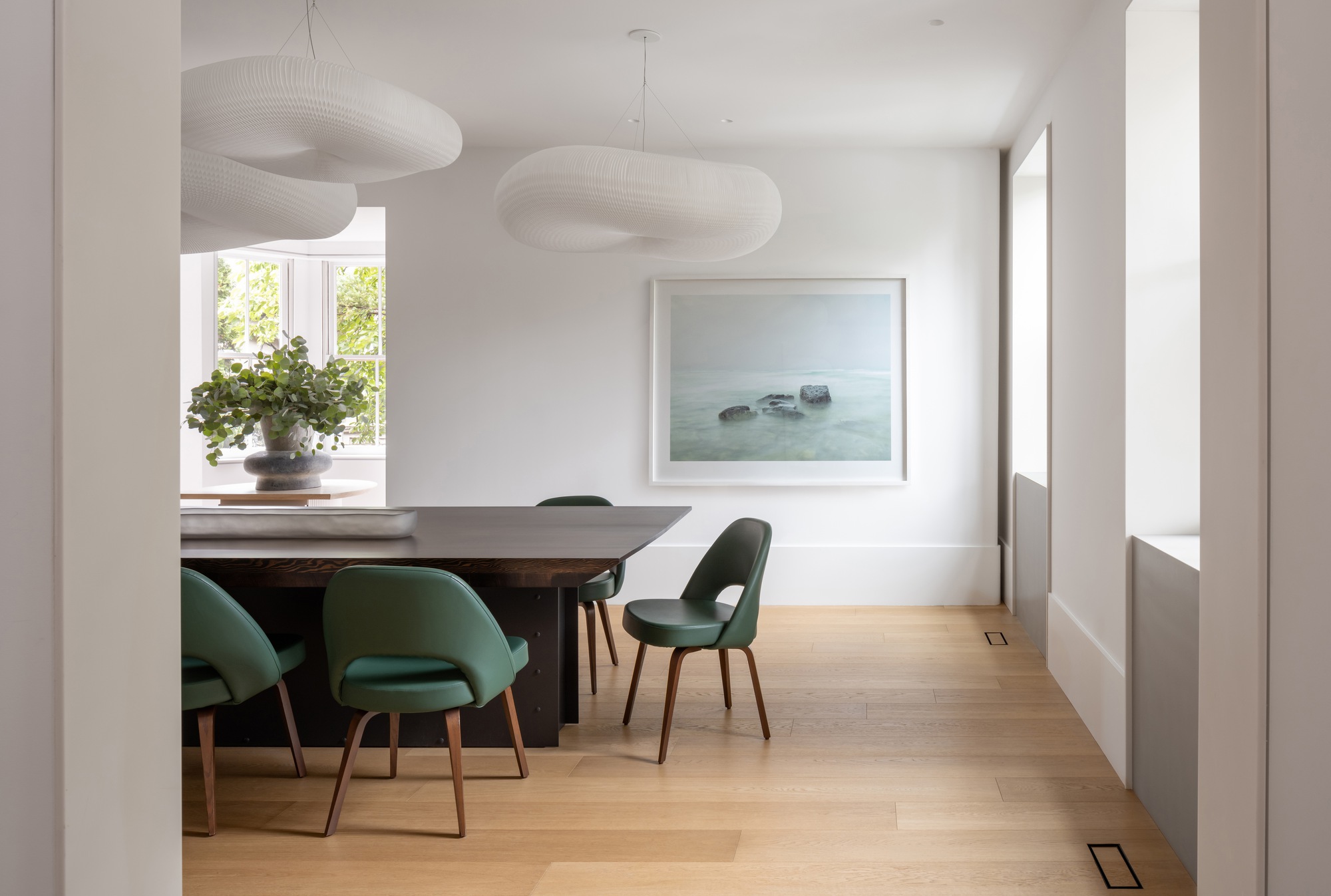Dining area with green leather chairs, a dark table and two cloud-like pendant lamps beside a bay window with garden foliage