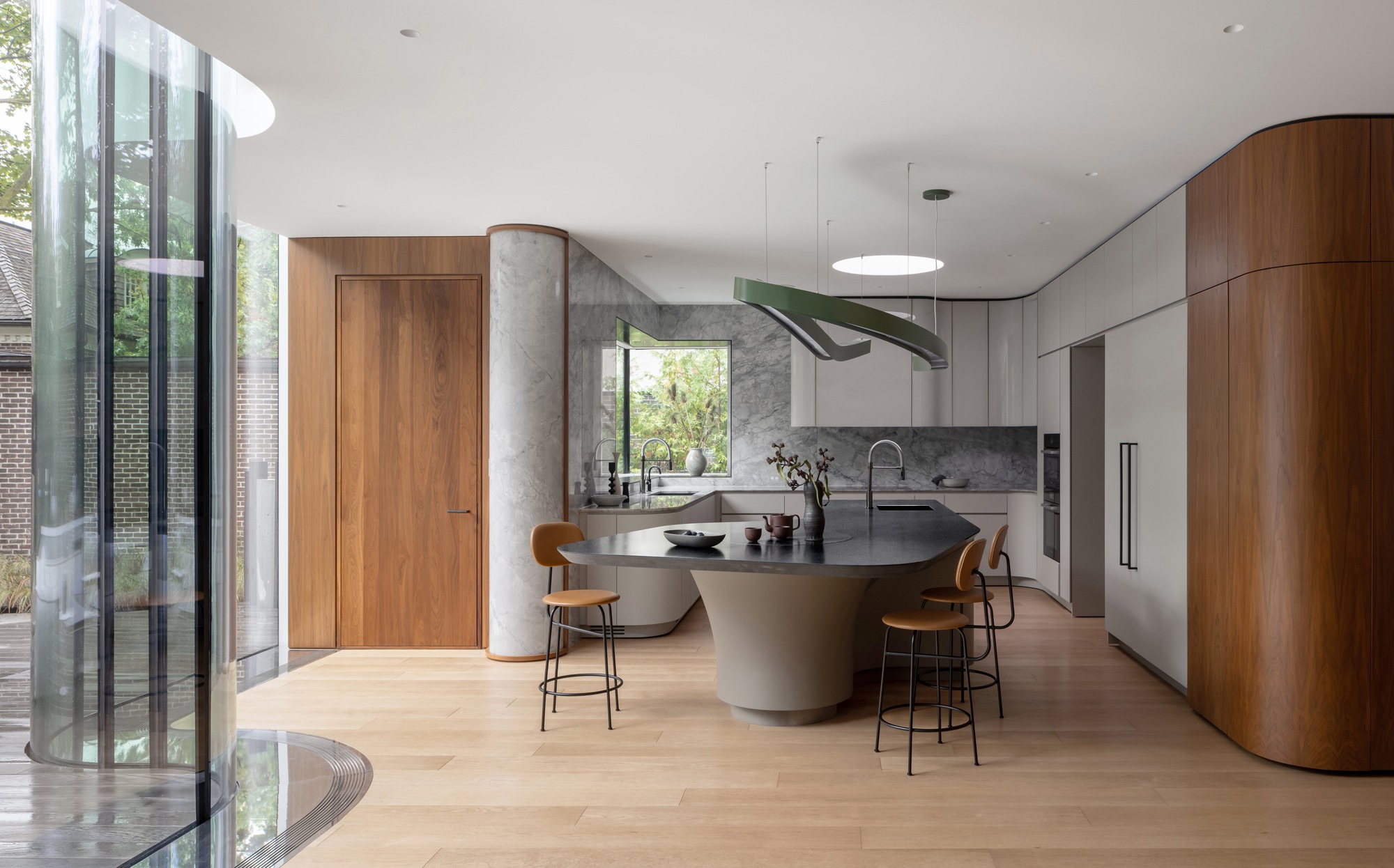 Kitchen with a curved walnut cabinet wall, grey marble column and island with leather stools, beside a curving glass wall facing a brick garden
