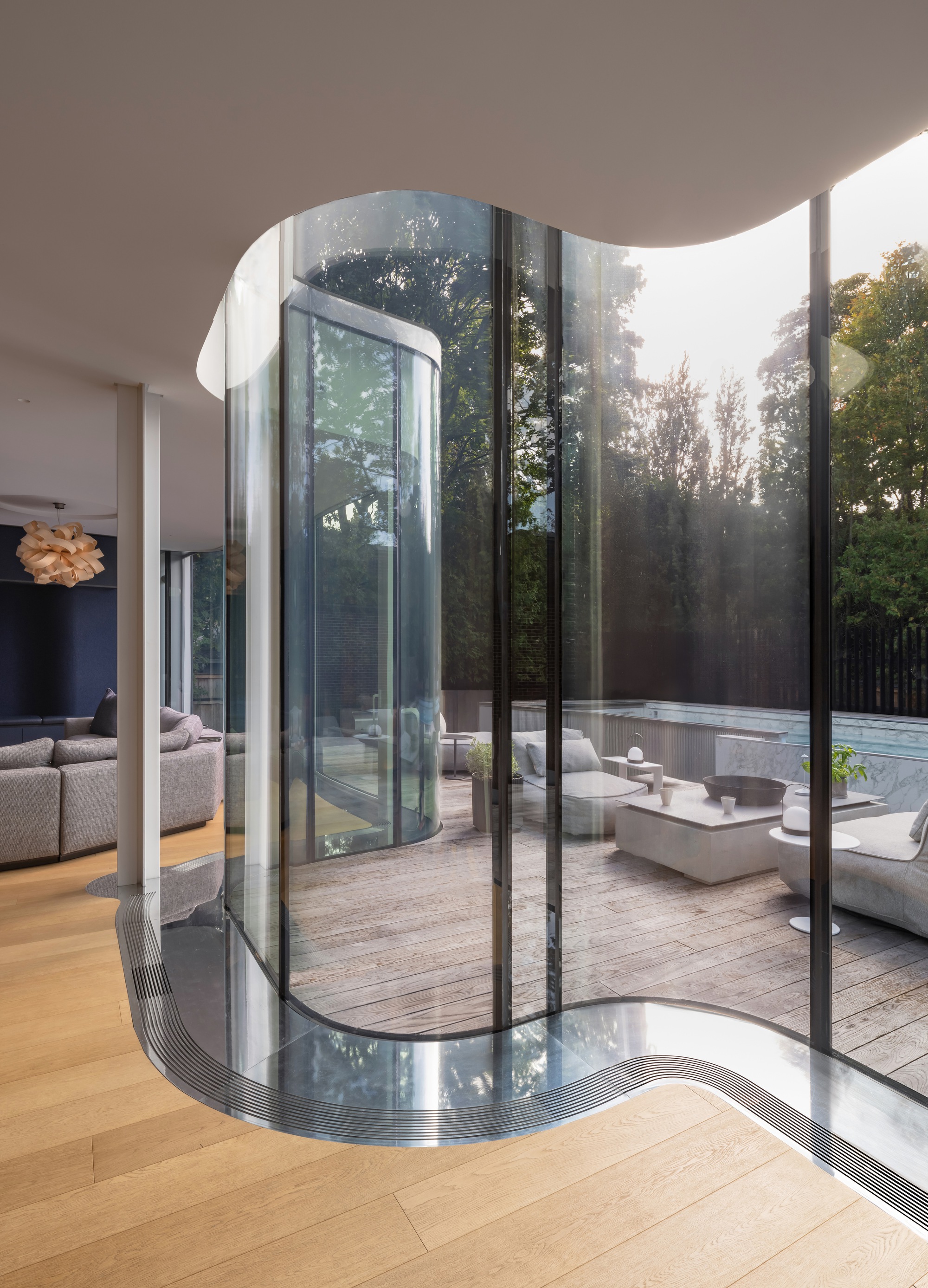 Curved glass wall with a metal floor channel opening onto a timber terrace with lounge seating, with sunlit trees behind