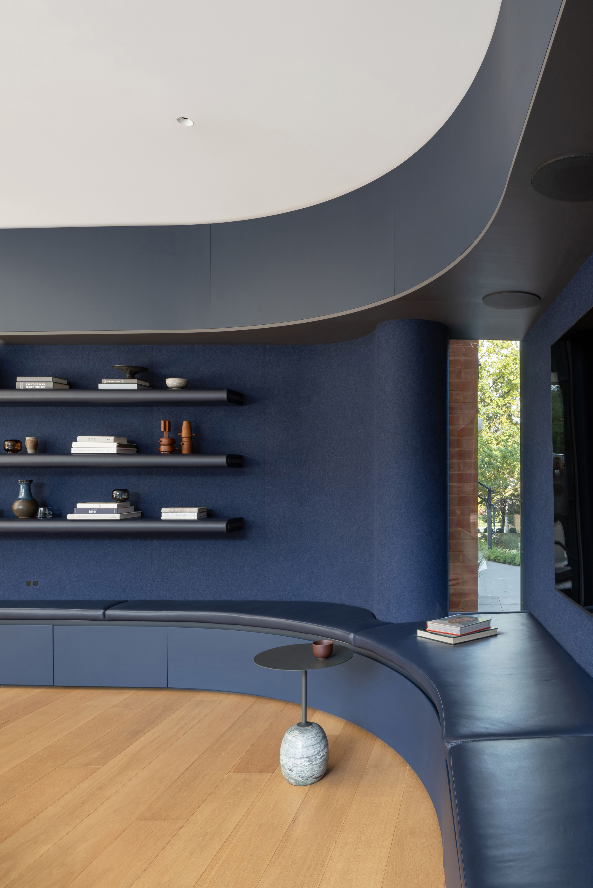 Curved blue felt wall with a leather bench, black floating shelves and a small marble-based side table, with a brick window slit letting in green daylight