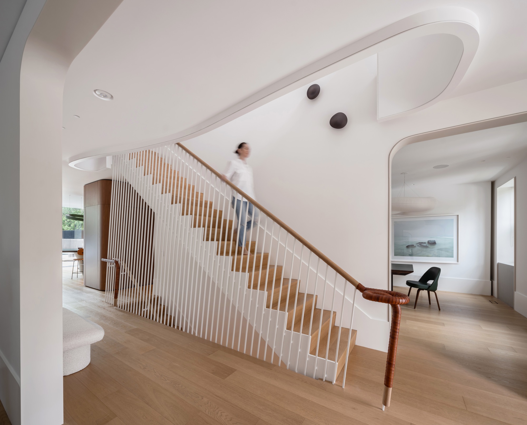 Open stair with white vertical balusters and oak treads, a leather-wrapped handrail, and a blurred figure descending past a curved ceiling edge