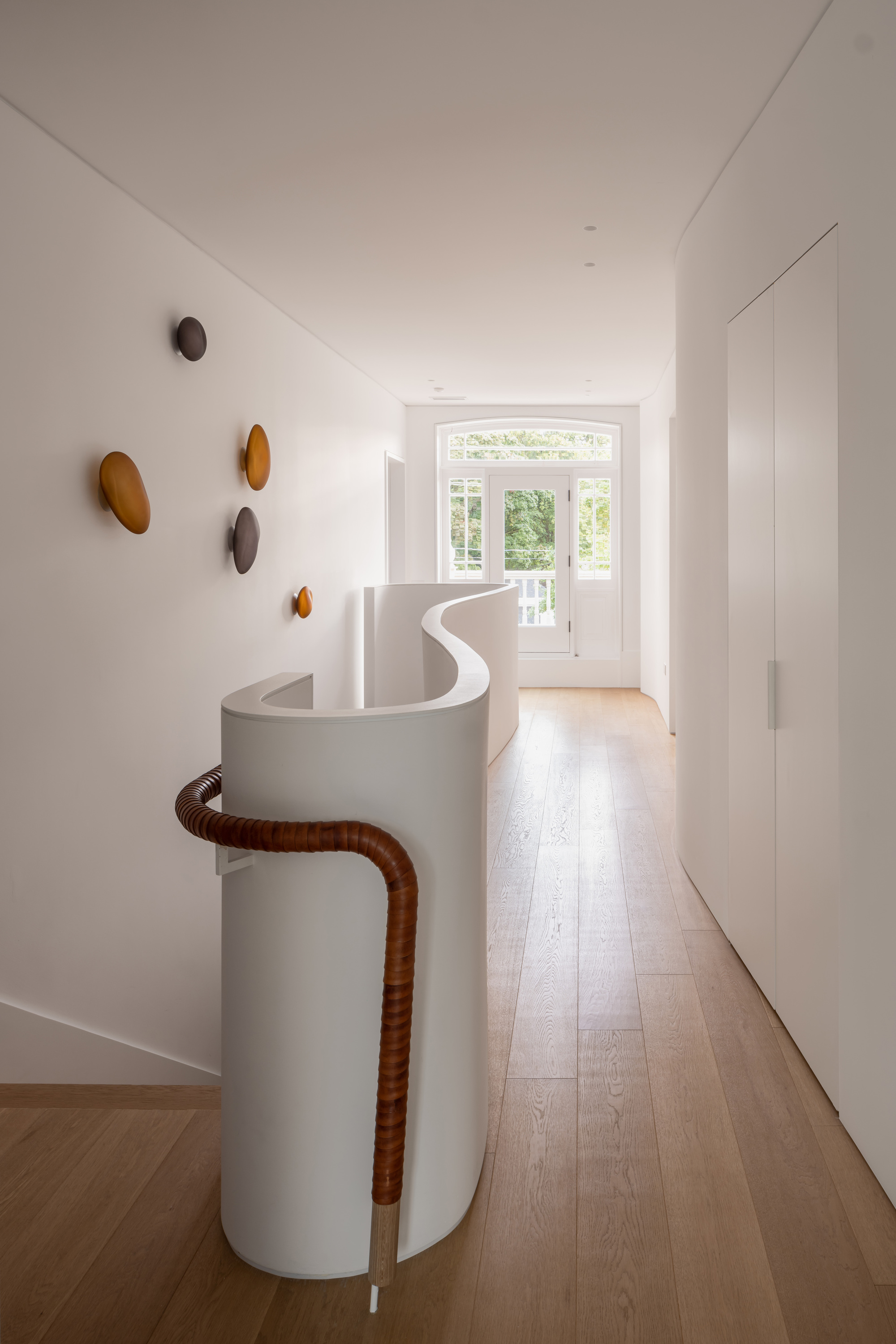 Upper landing with a curved white balustrade, leather-wrapped rail and amber wall pegs, leading toward an arched window with garden greenery