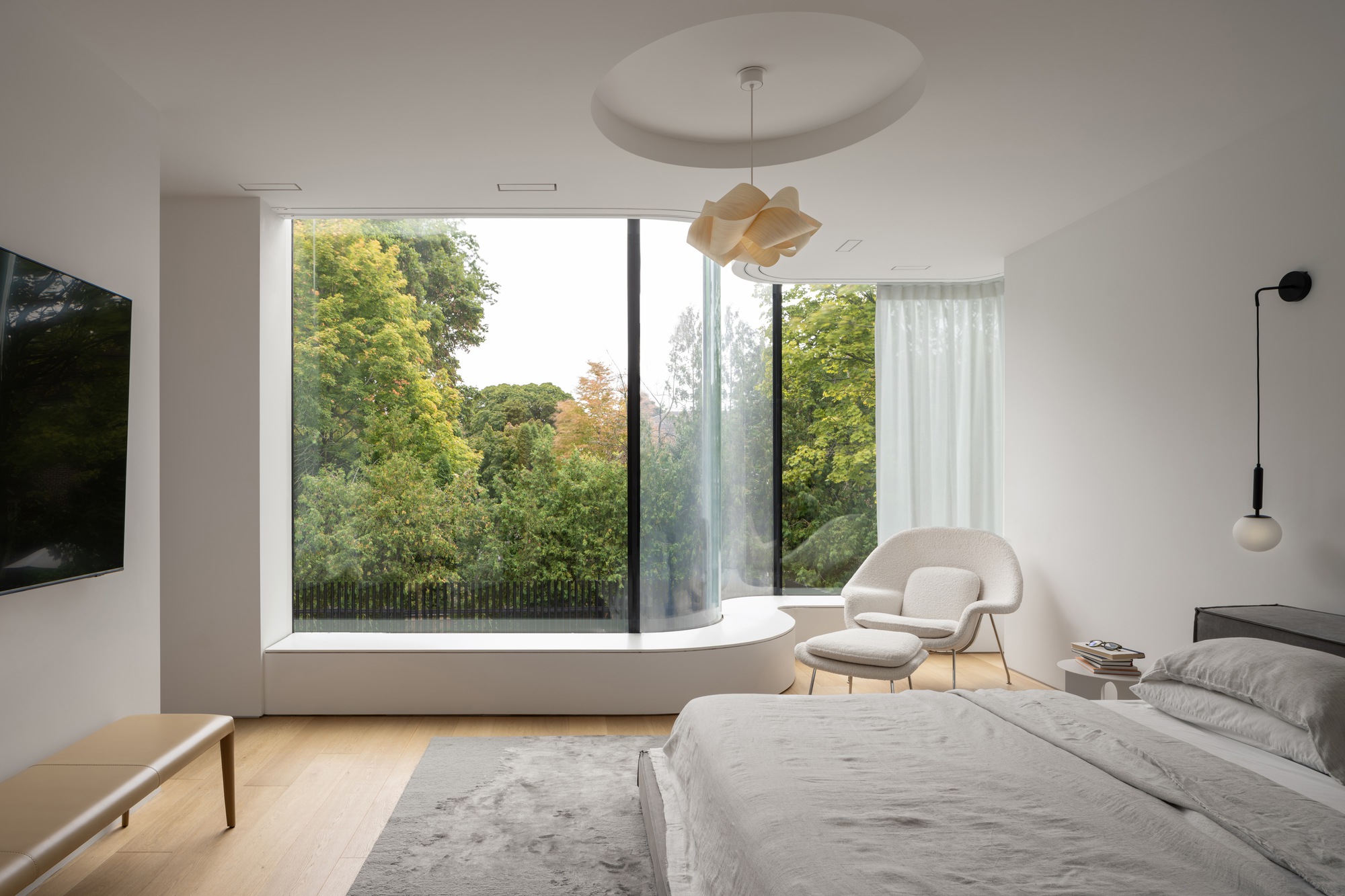 Bedroom with a curved glass corner and built-in window seat framing dense green trees, with a white lounge chair beside the bed