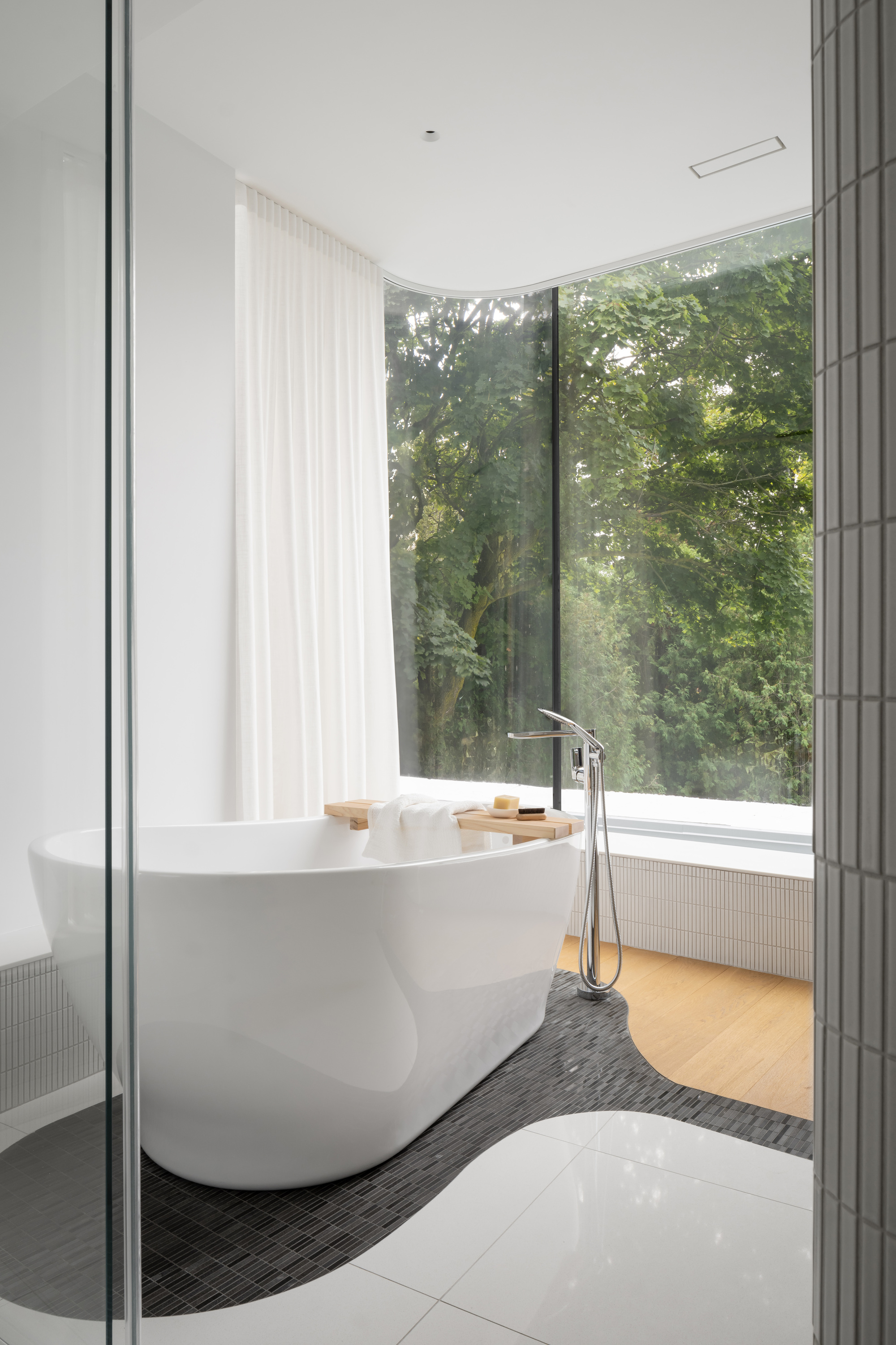 Freestanding white tub on a curving black mosaic floor inset, set before a corner window with tall trees and sheer curtains