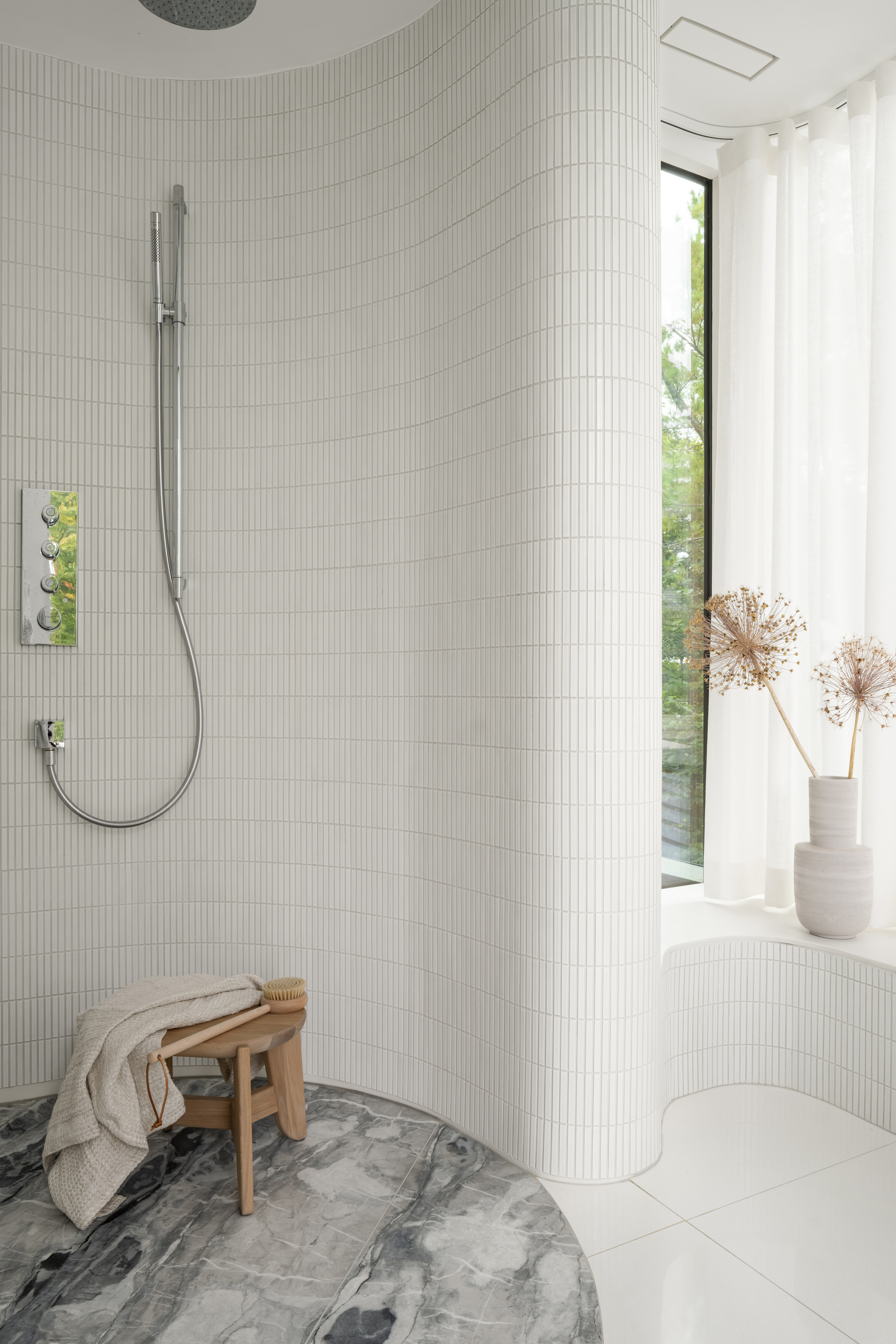 Curved shower wall clad in narrow white tiles, with a wooden stool on grey marble and dried allium stems by the window