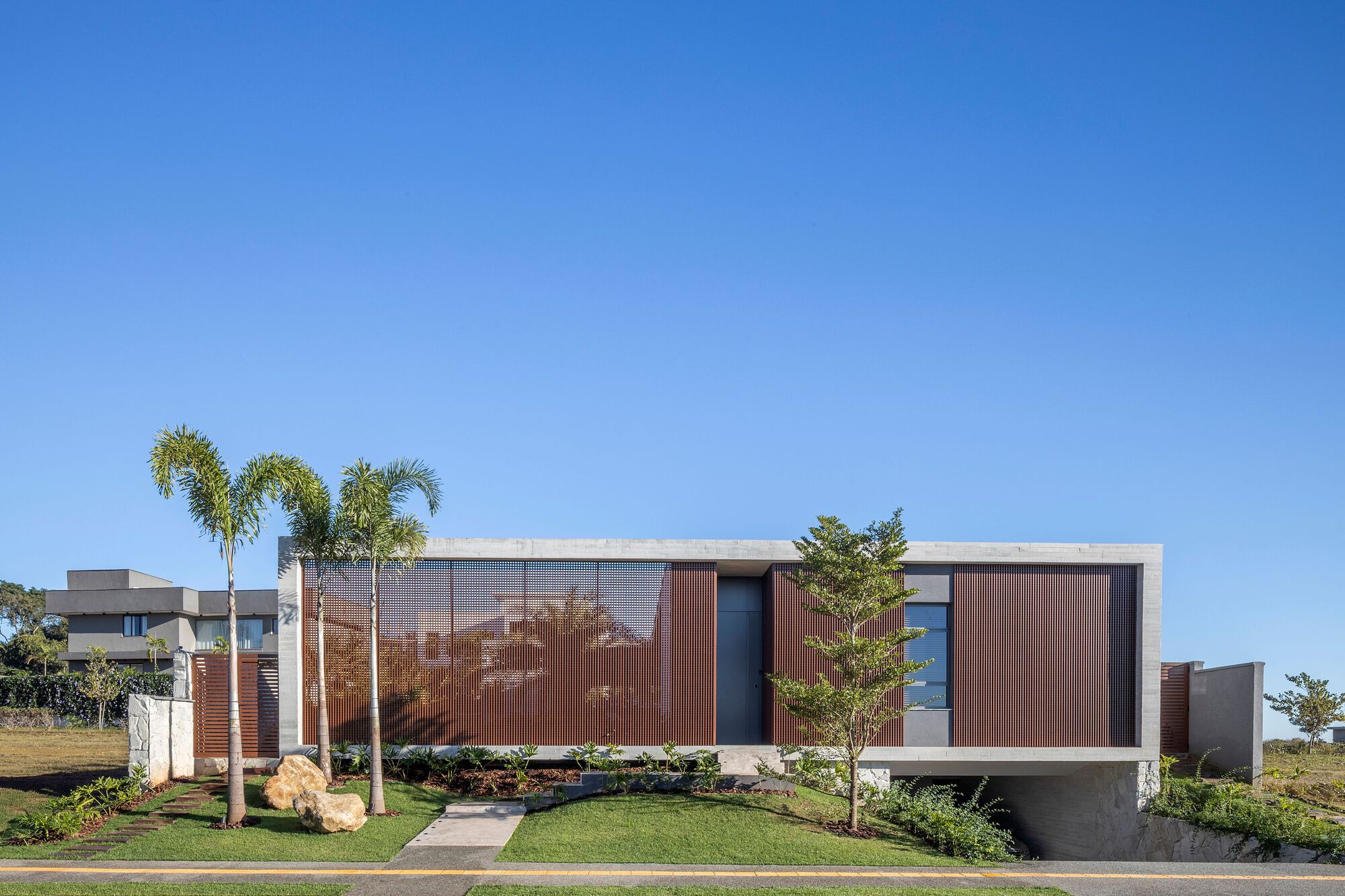 Street facade of a low concrete-framed house with timber slat panels, palms and a young tree under a clear blue sky