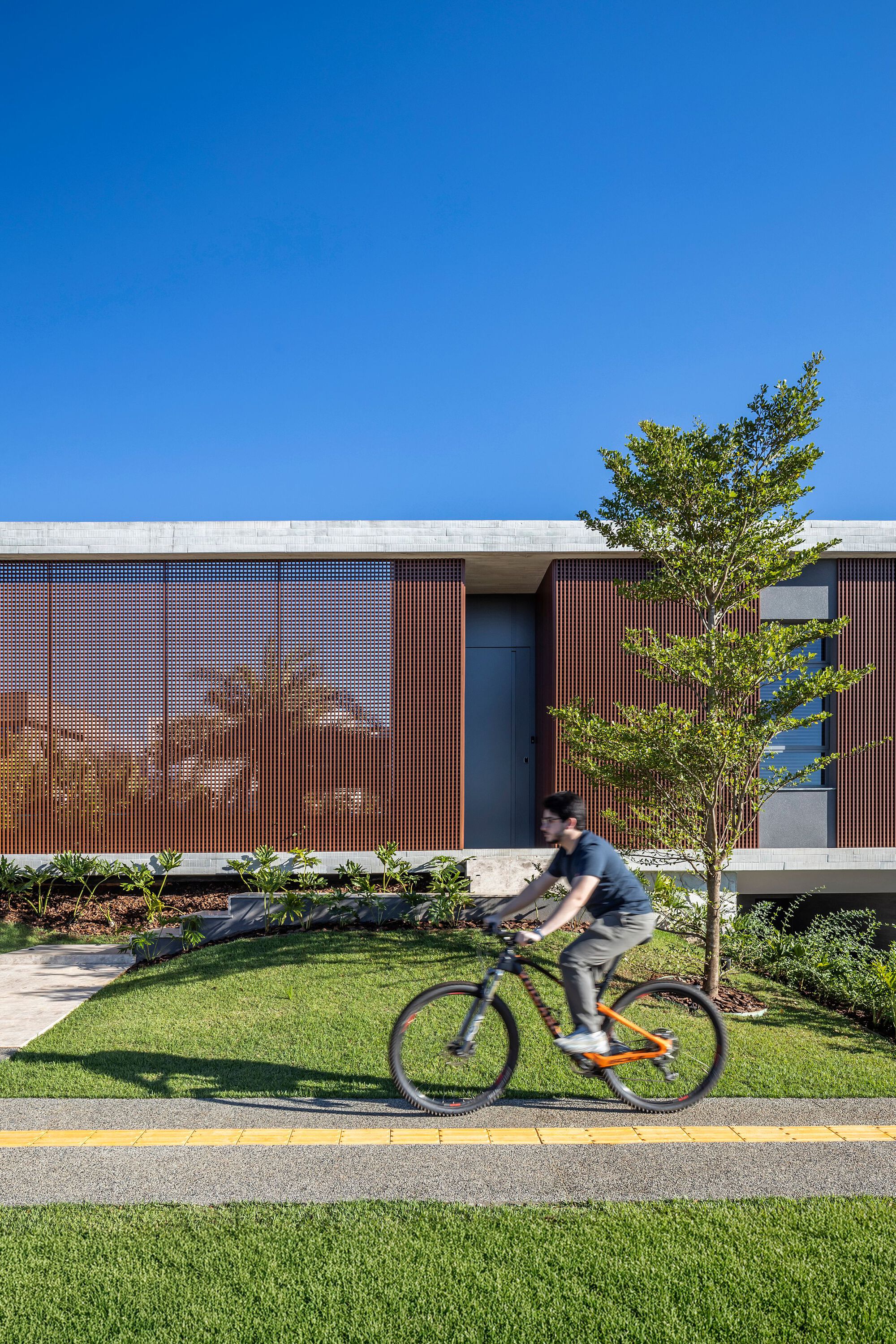 Street-side facade of rust-toned timber slats and a concrete roof edge, with a cyclist passing on the path under clear blue sky