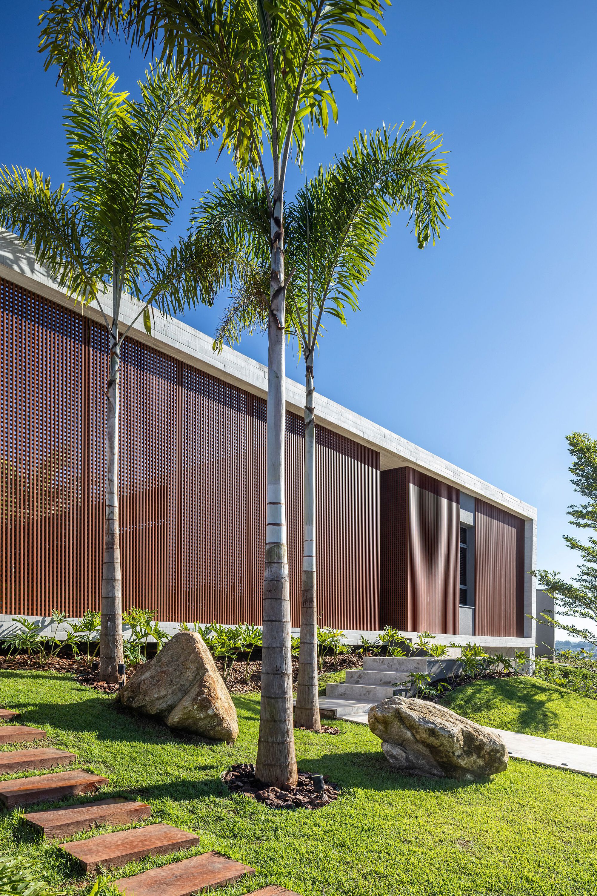 Facade of vertical timber screens behind slim palms and two boulders on a lawn under a clear blue sky