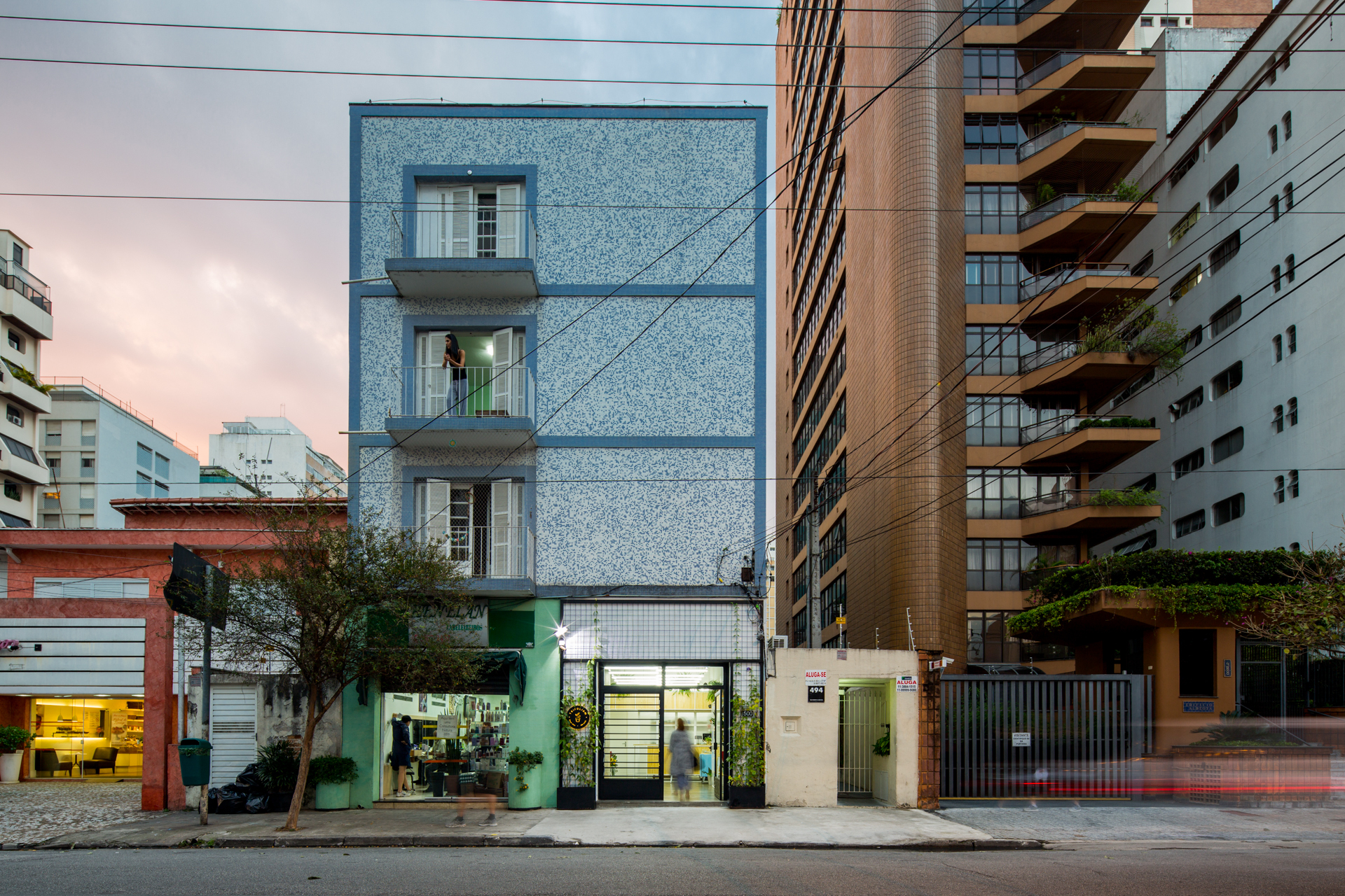 Street view of a narrow speckled blue-tiled facade with three small balconies, a shopfront below and a brick tower beside it at dusk