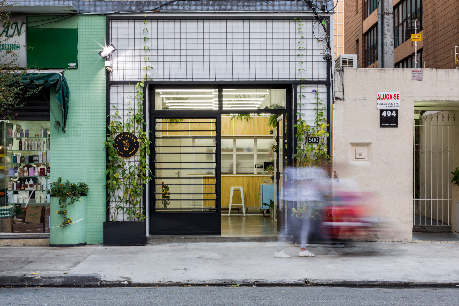 Shopfront with a black steel-framed glass door, a white tile grid and climbing vines, with a blurred pedestrian passing on the sidewalk
