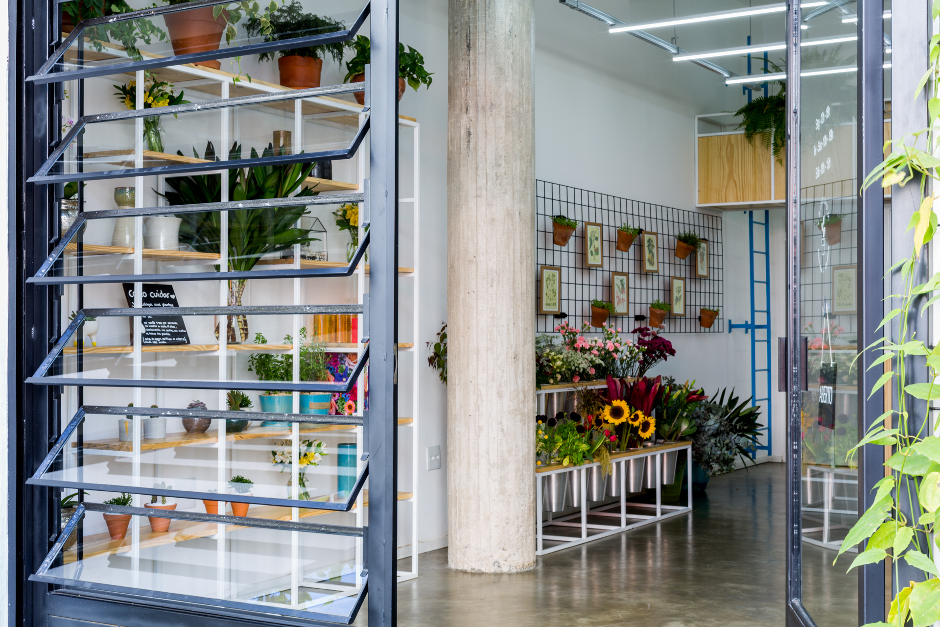 View through open louvered glass panes into a flower shop with a concrete column, wire wall grid and sunflowers on a steel stand