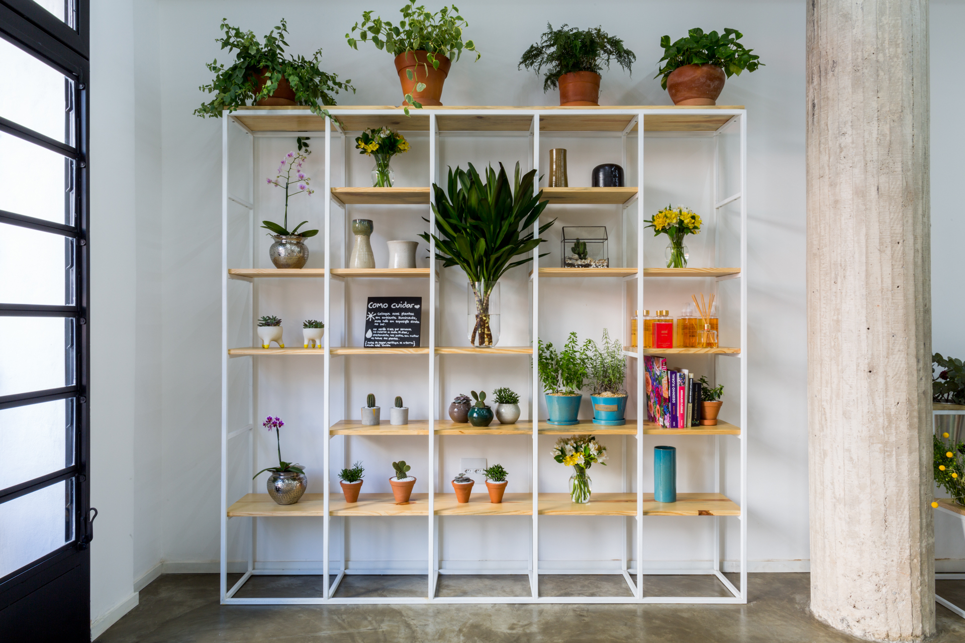 White steel shelving unit with timber boards holding potted plants, vases and books beside a board-formed concrete column in soft daylight