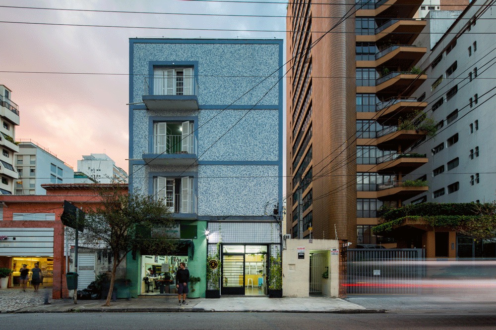 Street facade of a narrow blue speckled-tile building with small balconies and a shopfront below, seen at dusk beneath power lines
