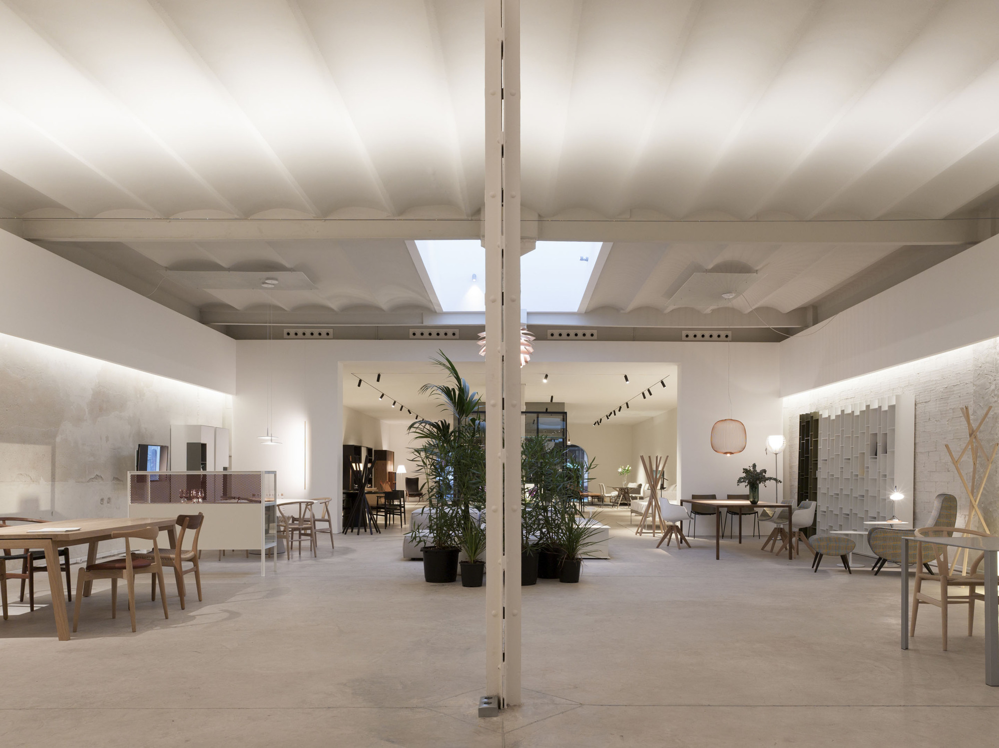 Open showroom hall with a central white column, skylight and potted palms, dining tables set along lit concrete walls