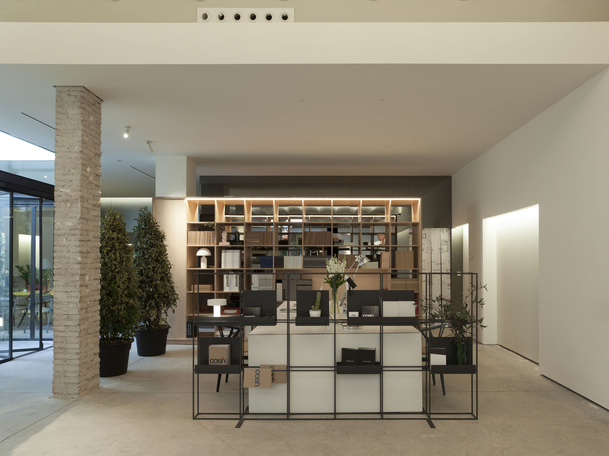 Timber shelving unit with a black steel grid display frame in front, beside a stacked brick column and clipped potted trees