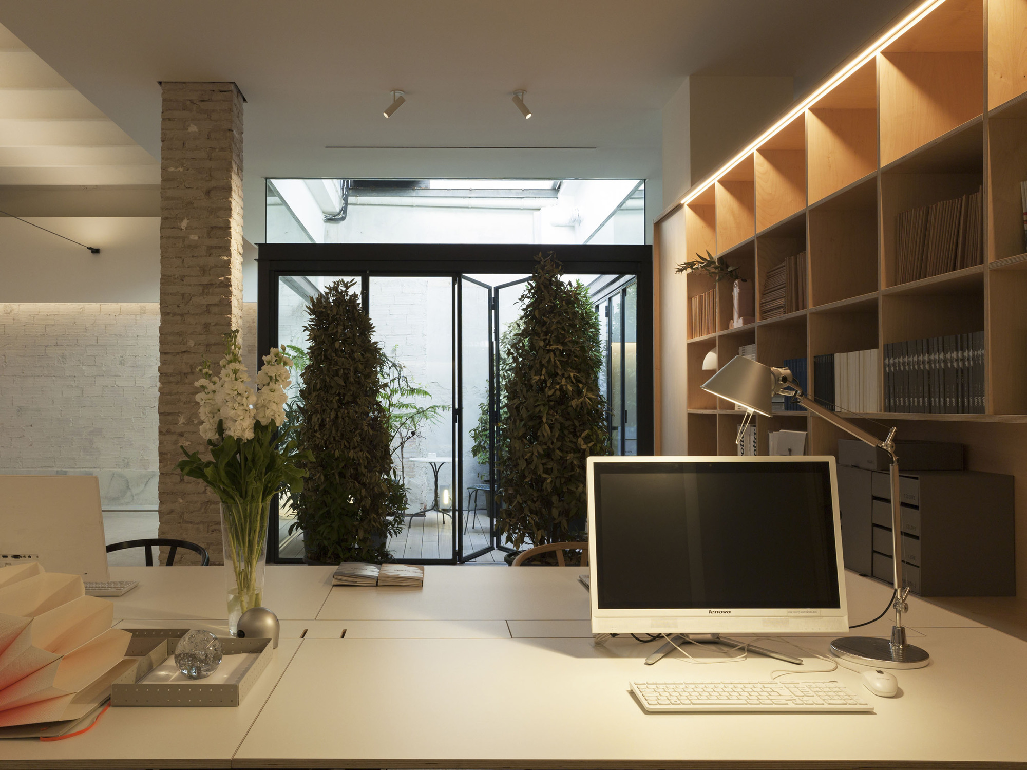Desk with monitor and task lamp facing a glazed folding door, where two conical shrubs stand in a daylit light well