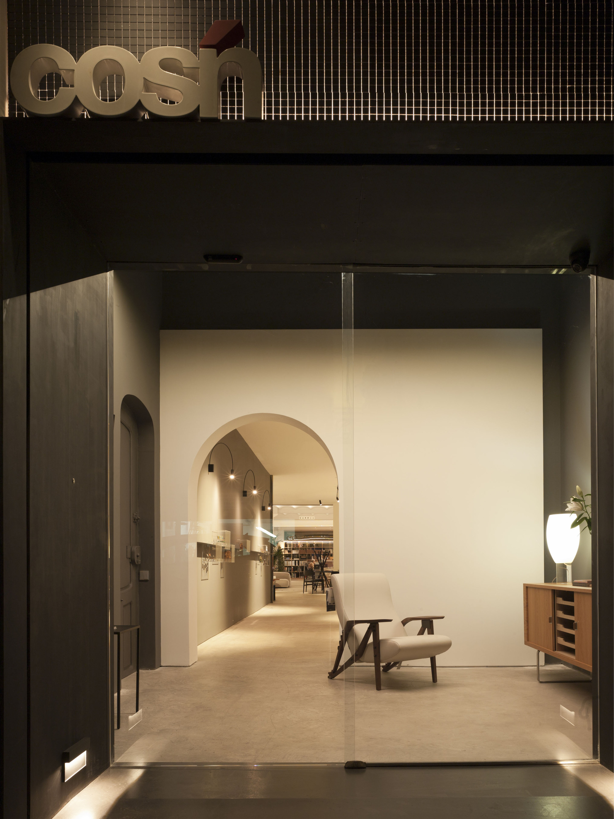 Night view through the glass entrance to an arched passage and a lounge chair, with a lettered sign above