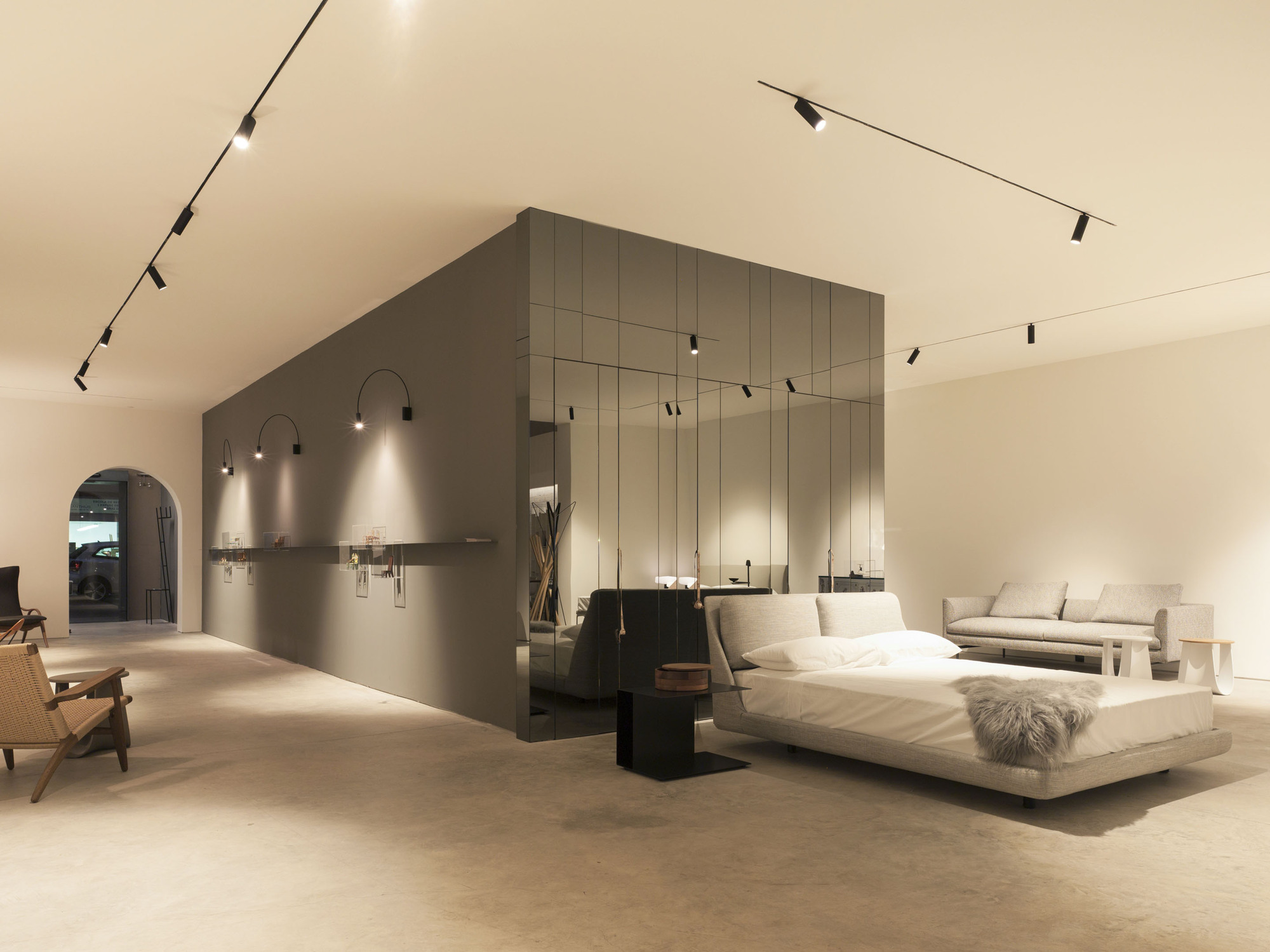 Mirrored glass volume beside a grey wall with arched sconces, a low bed in the foreground under black track lighting