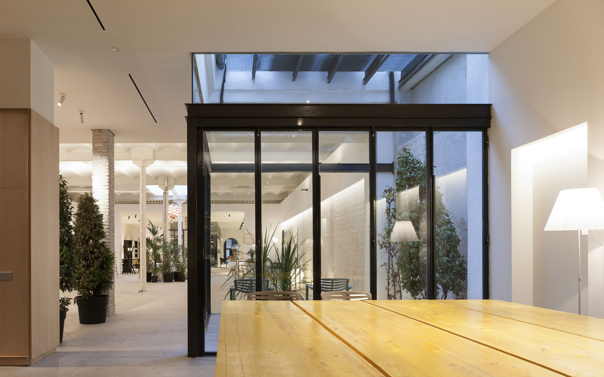 Black steel-framed glass enclosure with potted plants and a long pine table in an evening-lit open interior with brick column