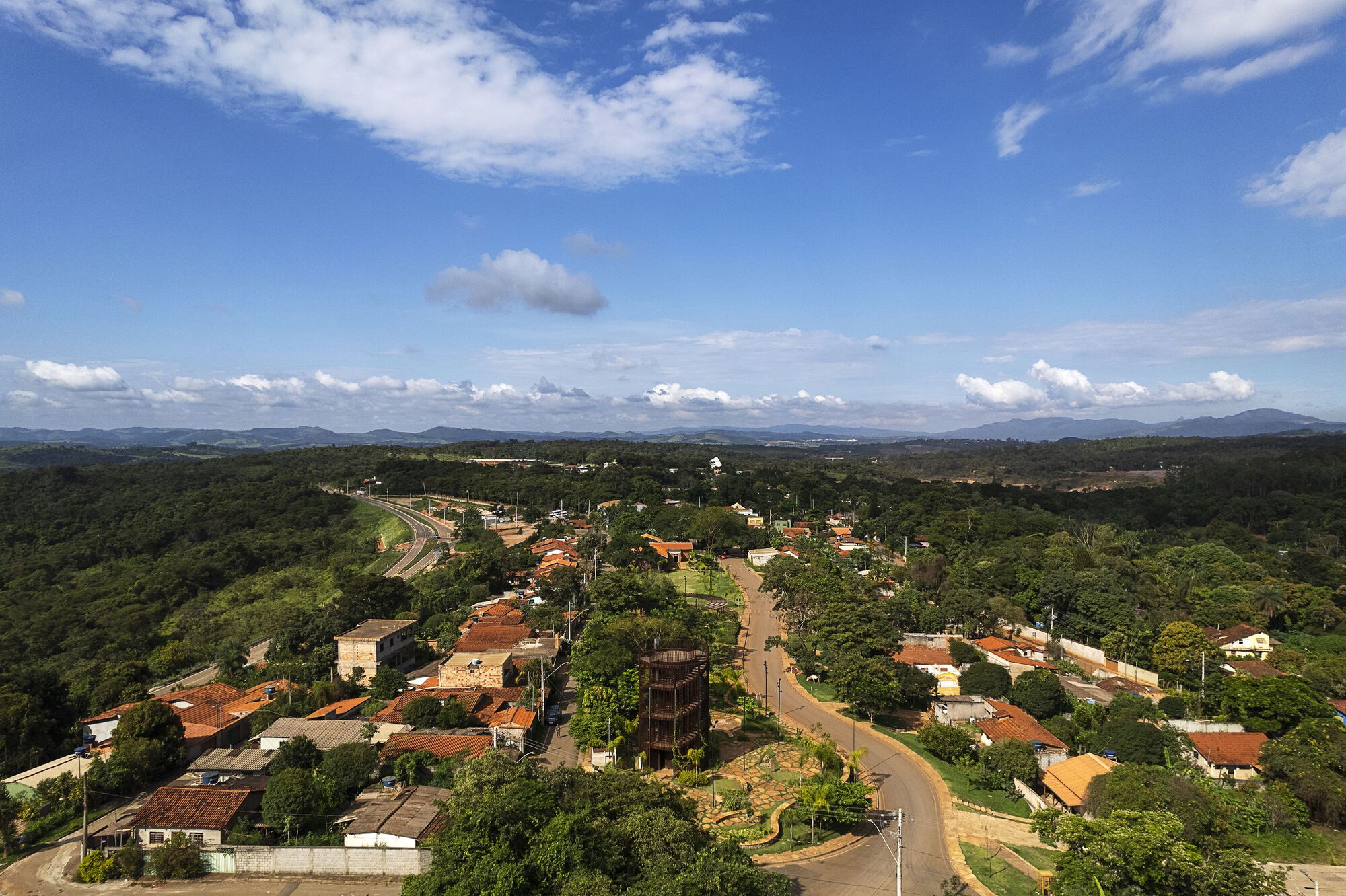 Aerial view of a small town with red-tiled roofs, a curving road and a slatted tower among trees under scattered clouds
