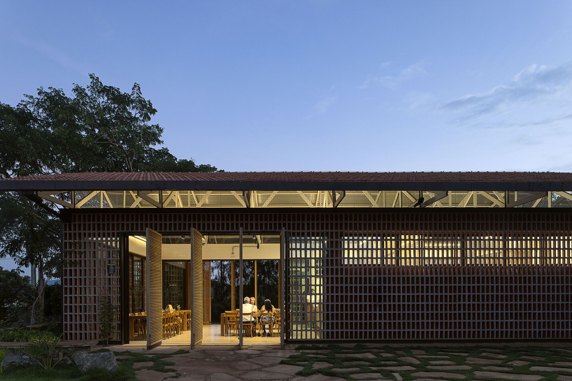 Brick lattice pavilion with pivoting wooden doors open, showing diners at long tables beneath a clerestory at blue dusk