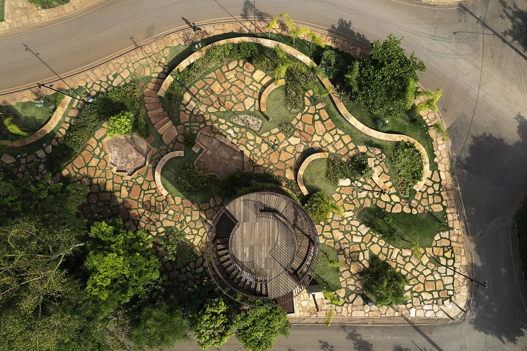 Top-down view of a stone-paved garden with winding low walls and planting beds, circling the round tower roof beside a road