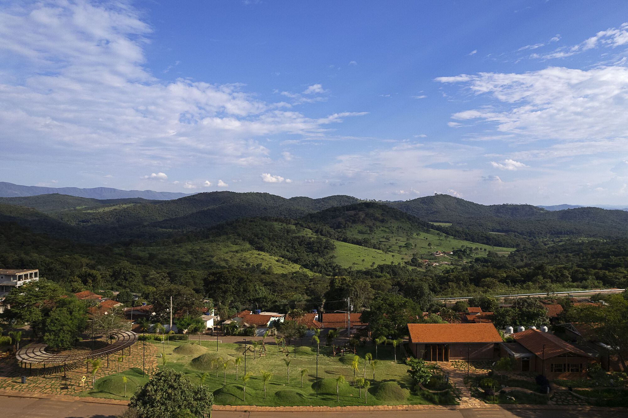 Elevated view of a village square with grassy mounds and terracotta-roofed buildings below forested green hills under a bright blue sky