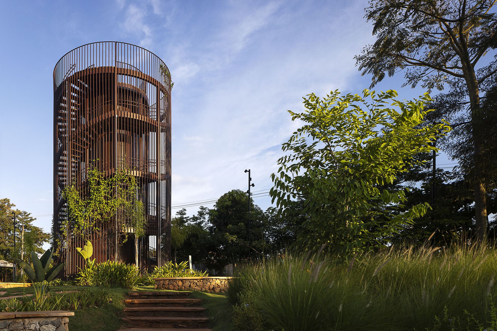 Cylindrical weathered steel tower with vertical slats and climbing vines, reached by stone-edged steps beside tall grasses in late light