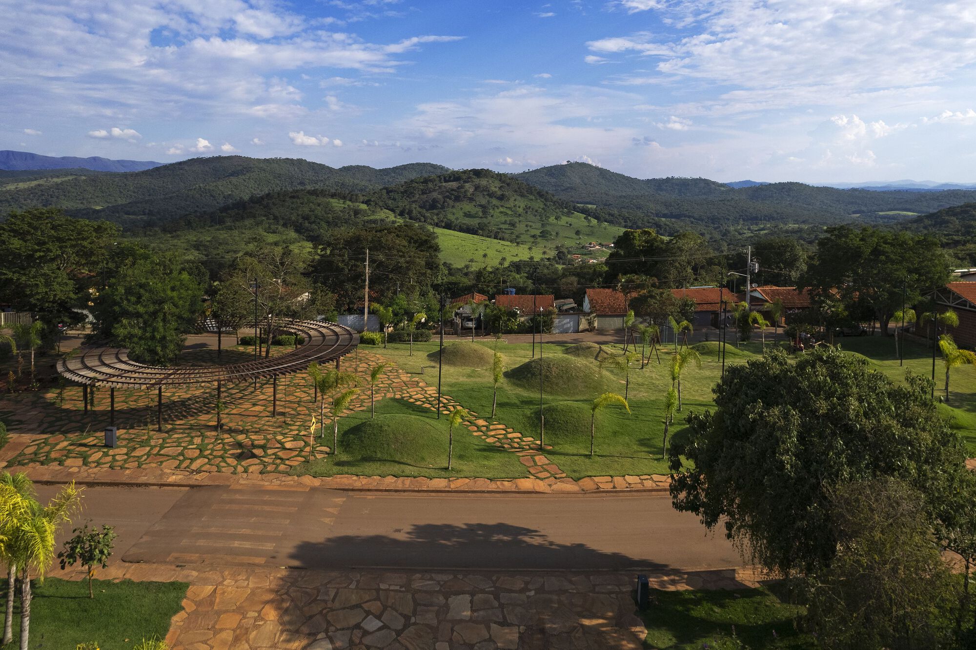 Aerial view of a curved timber pergola and stone-paved plaza beside grassy mounds planted with palms, hills rising behind