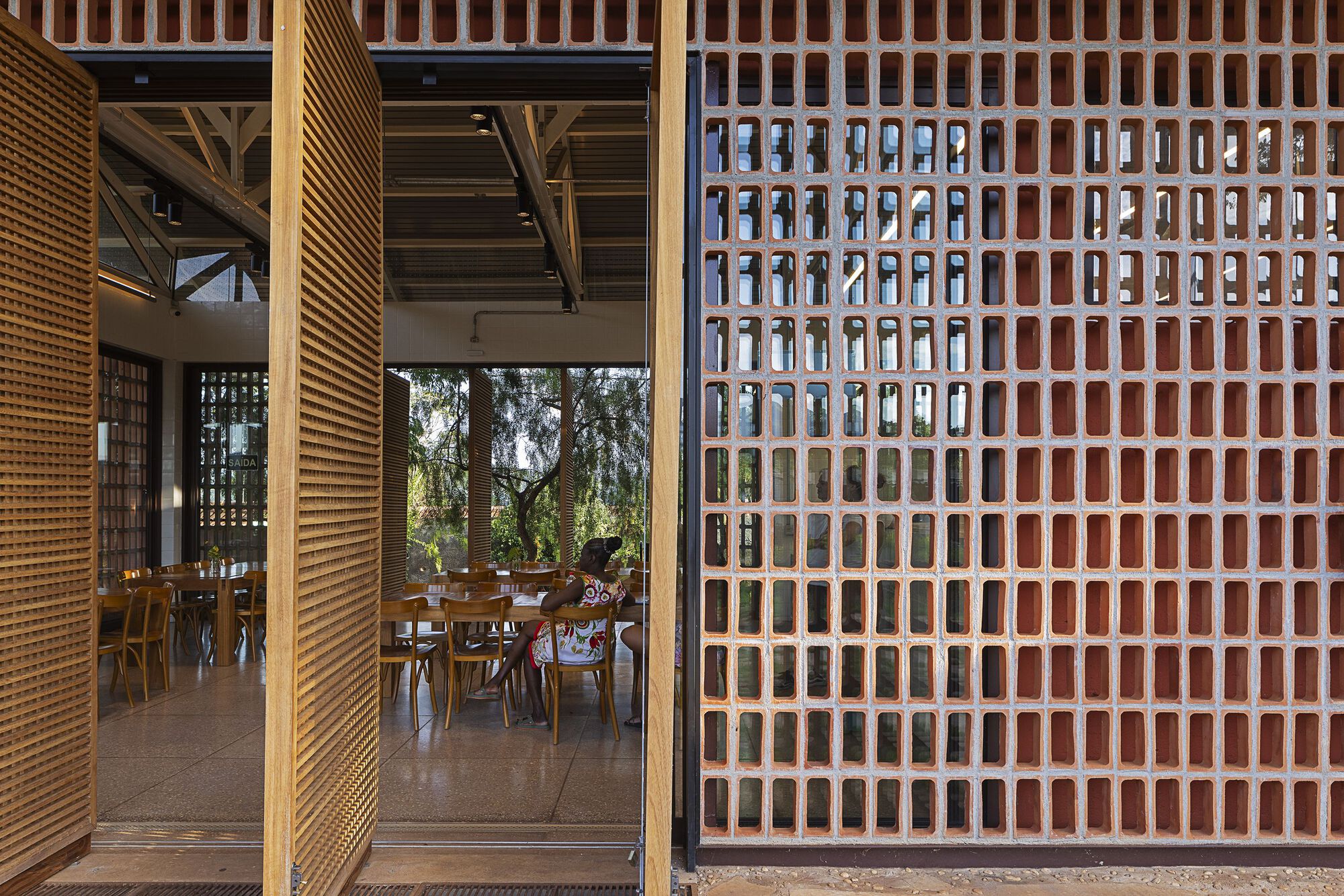 Pivoting timber slat doors open beside a perforated terracotta brick wall, revealing diners seated at long tables inside