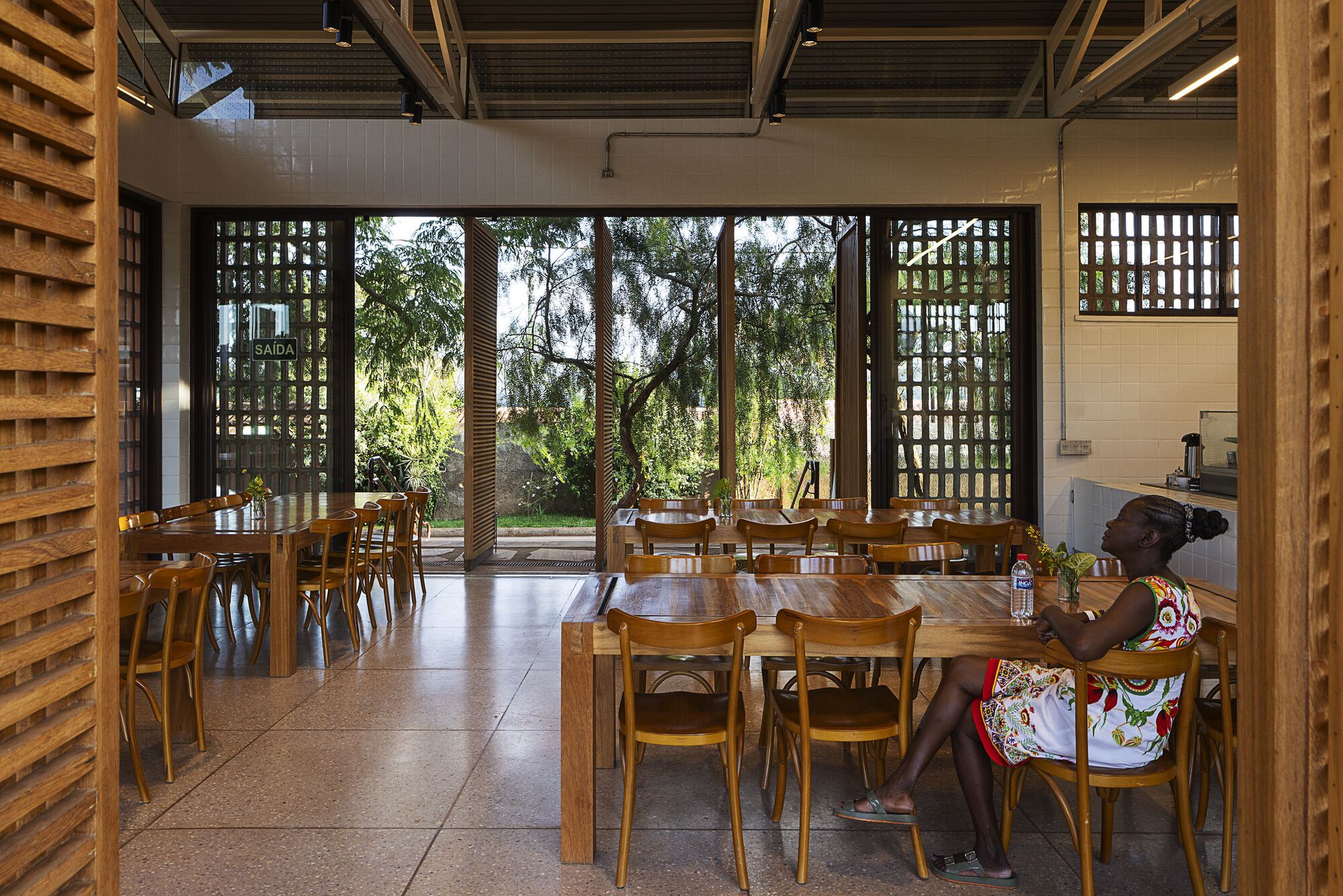 Dining hall with long timber tables and bentwood chairs opening through louvered doors to a tree-filled garden, a woman seated at right