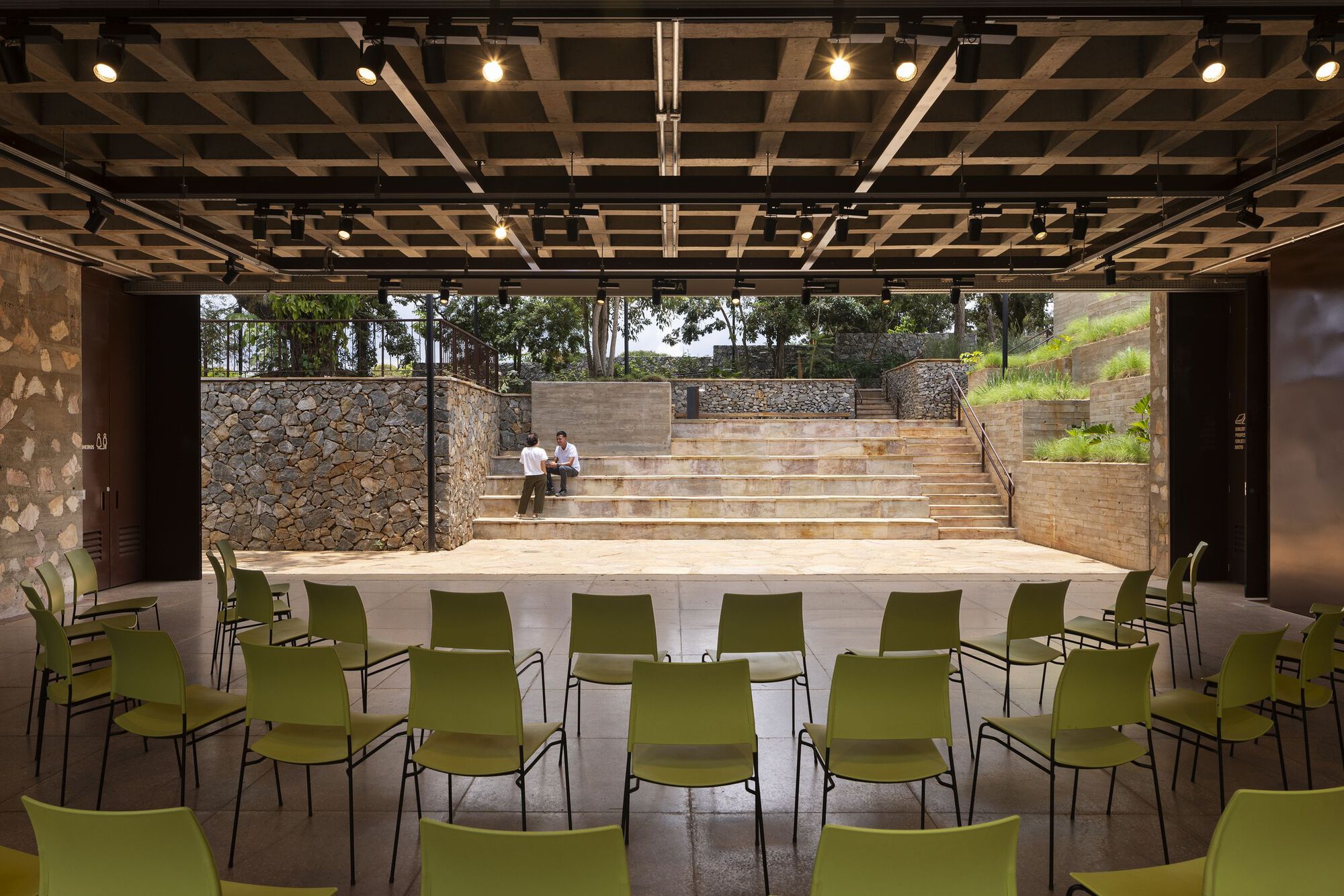 Covered hall with lime-green chairs facing a stepped stone amphitheater, two people sitting on the terraces under a gridded timber ceiling