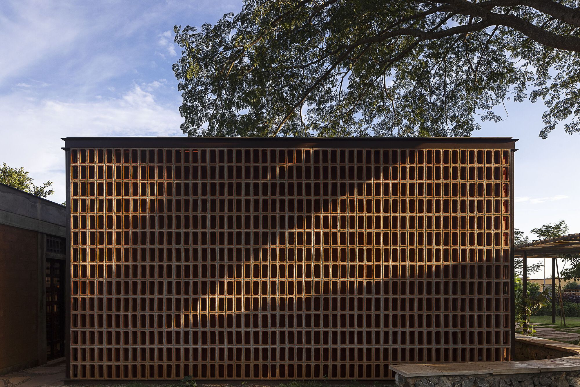 Freestanding perforated brick screen wall with diagonal tree shadow cast across it beneath a large canopy and scattered clouds