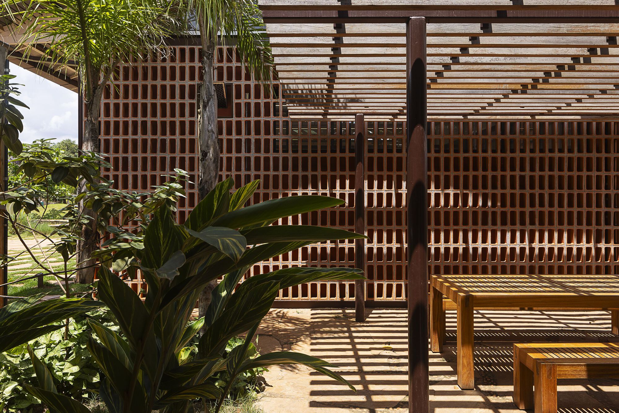 Wooden benches and tables in striped pergola shadow in front of a brick lattice wall, tropical leaves crowding the foreground