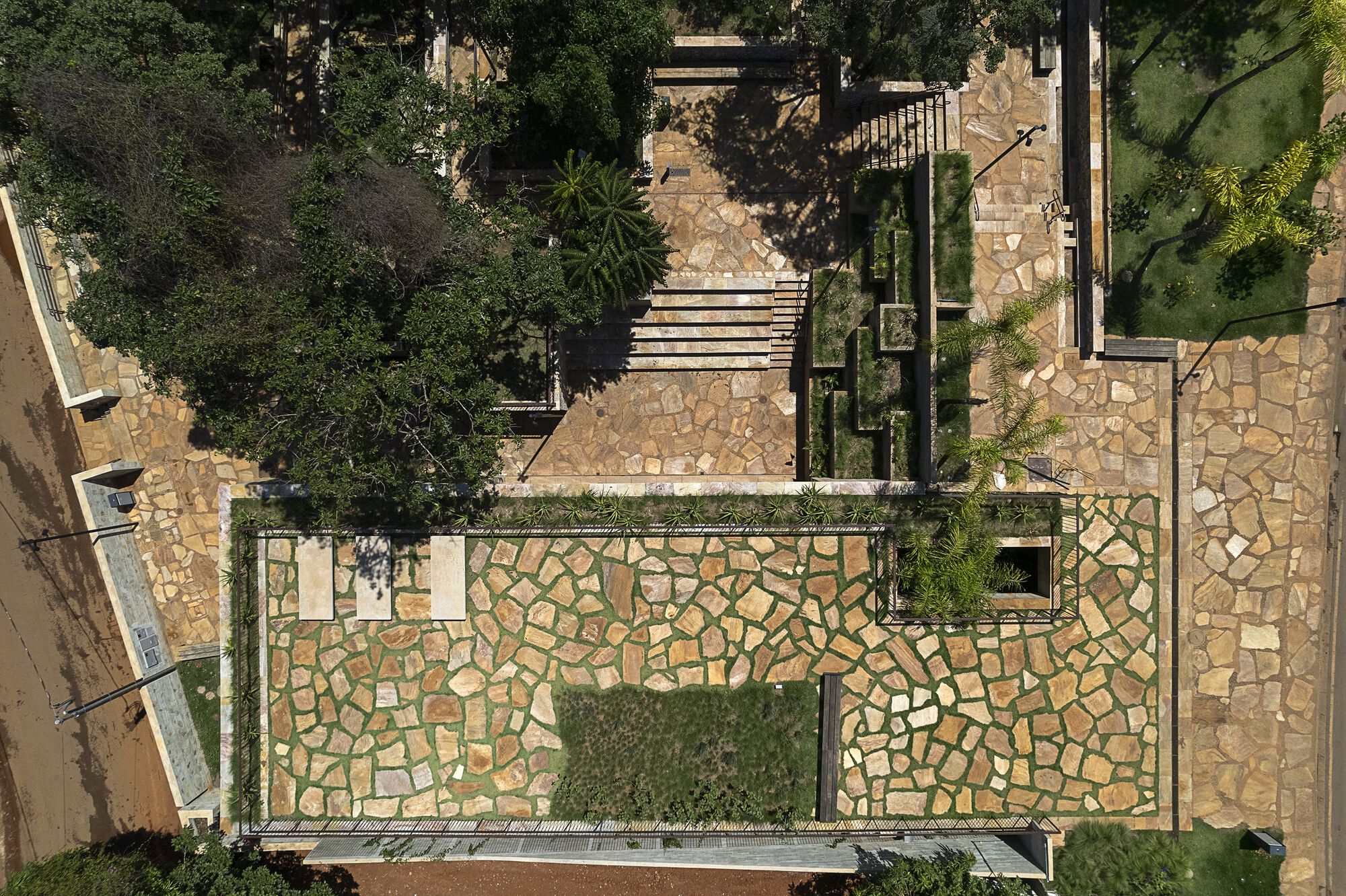 Drone view of a flagstone-paved rooftop with grass joints, a planted bed and stepped paths beside tree canopy