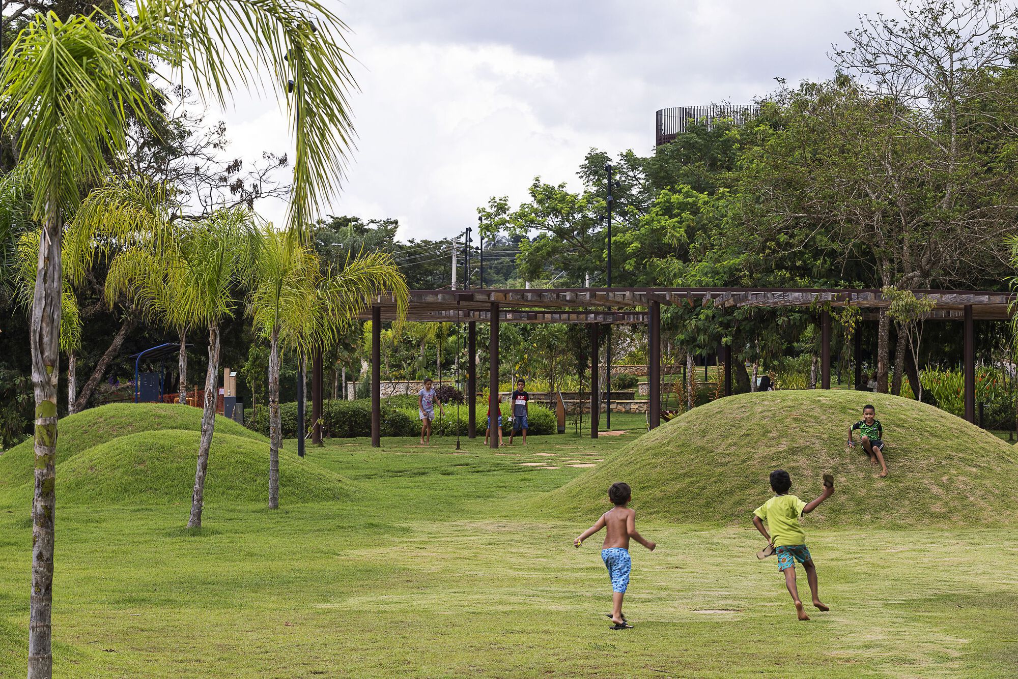 Children running across lawn toward grassy mounds and a timber pergola under a cloudy sky
