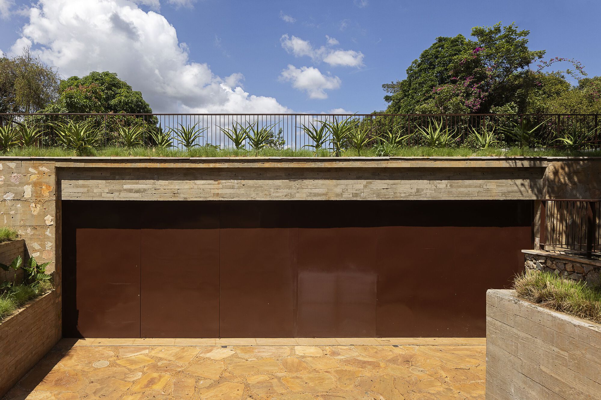 Rust-colored steel garage door beneath a stone-banded beam with agave plants and a railing above, under blue sky