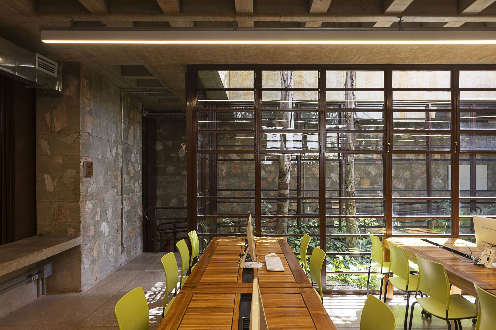 Computer desks with lime green chairs beside a timber-framed glass wall overlooking a planted courtyard and tree trunks