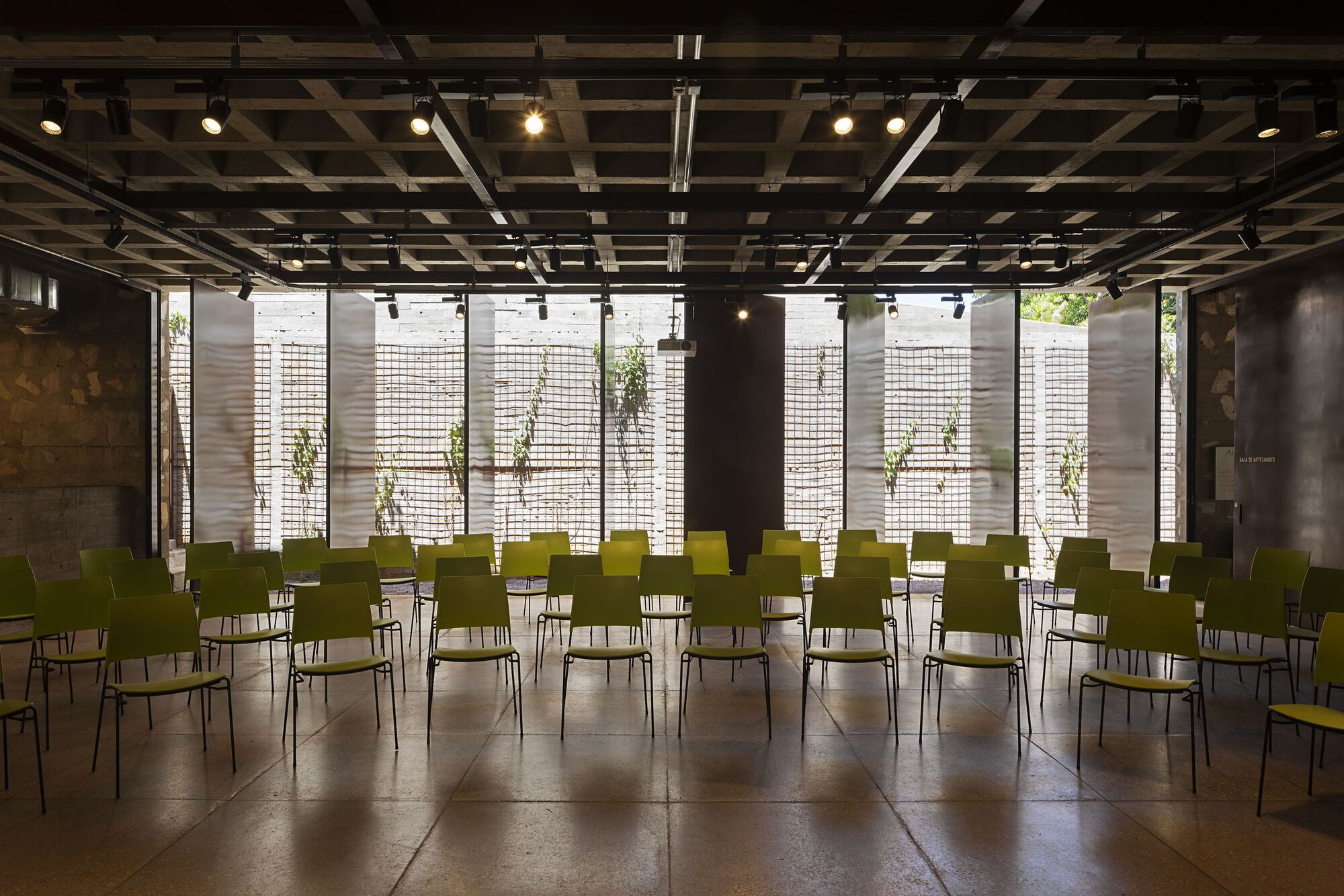 Lecture room with rows of green chairs under a coffered concrete ceiling and track lights, facing a screened garden wall